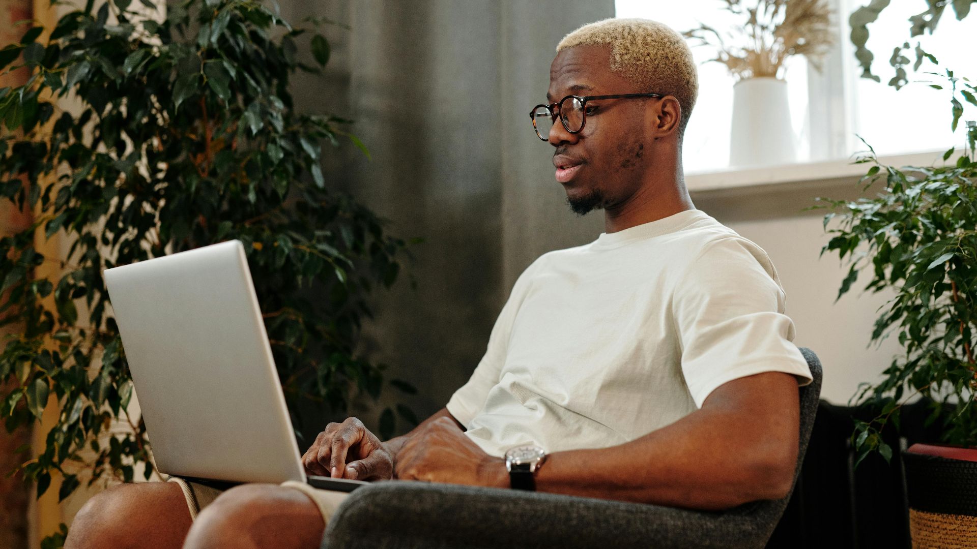 Focused young man with glasses working on a laptop in a cozy indoor space surrounded by plants.