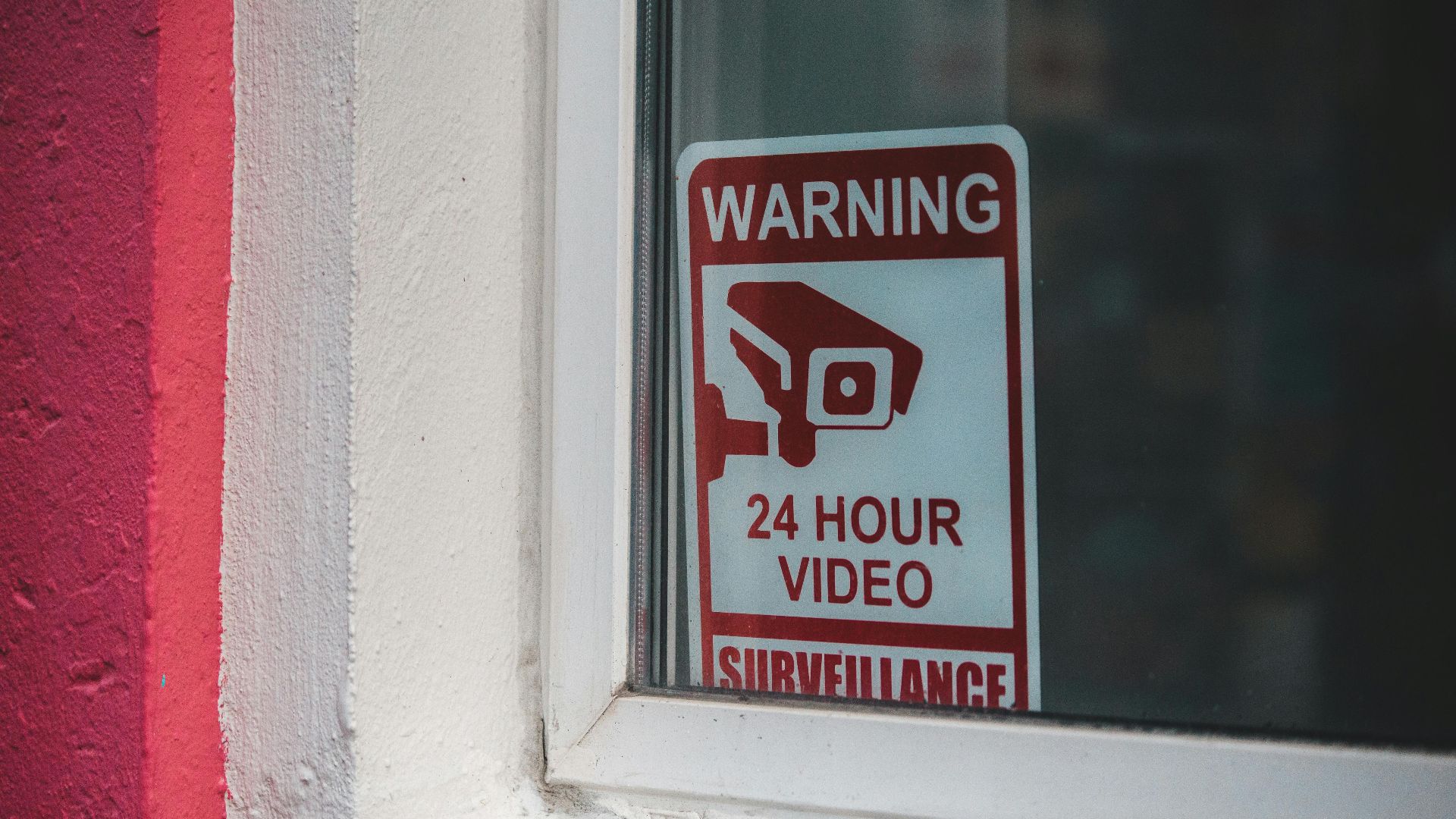 Red warning sign of video surveillance placed behind window of building in daytime