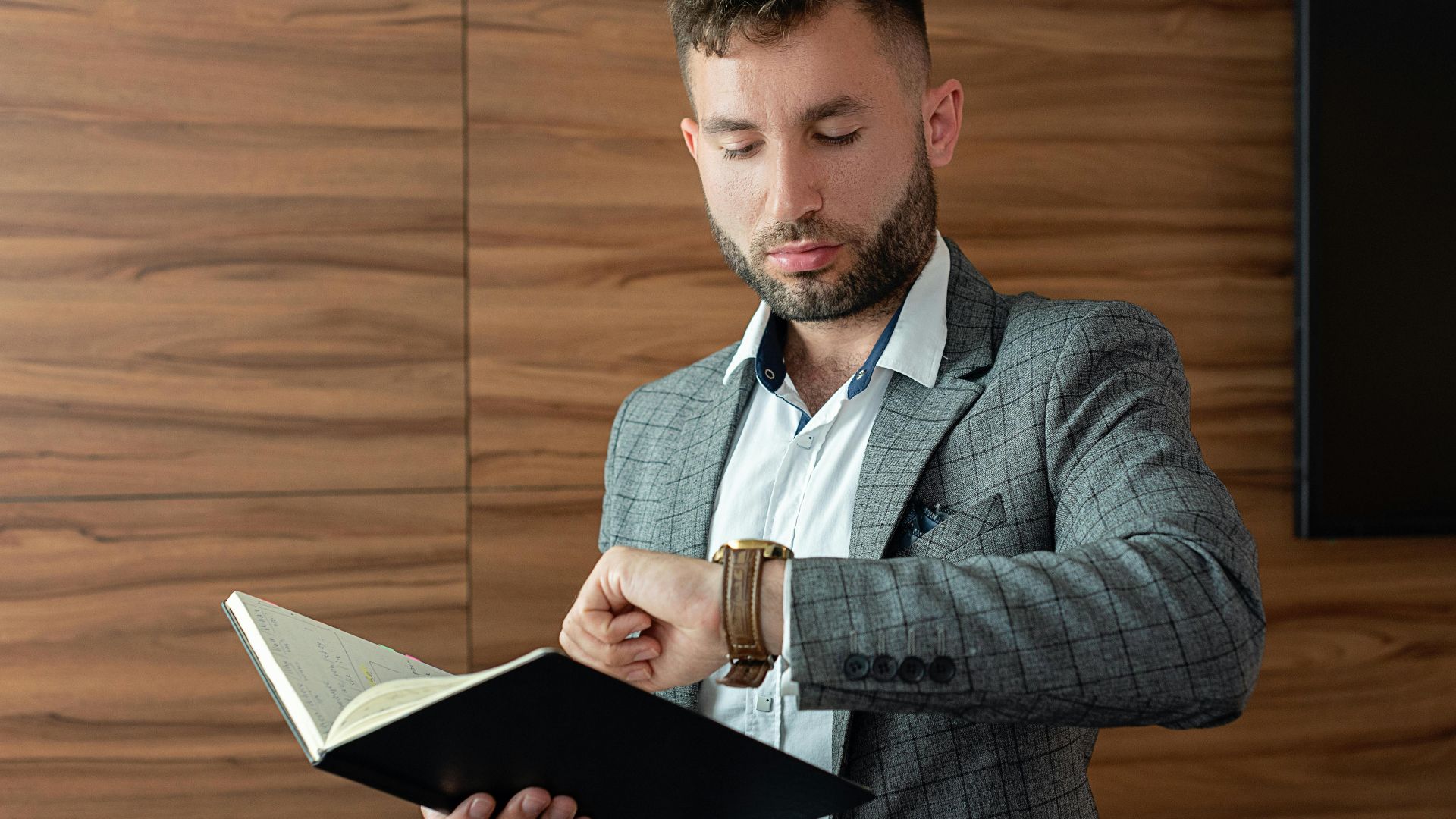Young businessman in gray suit checking wristwatch while holding a notebook indoors.