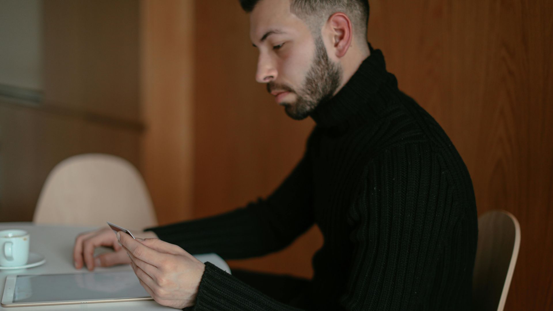 Focused man in a black sweater uses a digital tablet at an indoor table, emphasizing technology and concentration.