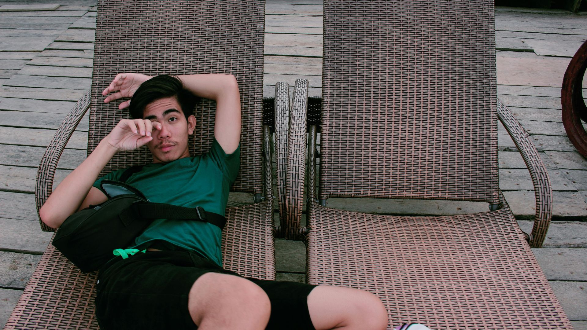 A young man relaxes on a wicker sunlounger on wooden planks, enjoying leisure time outdoors.