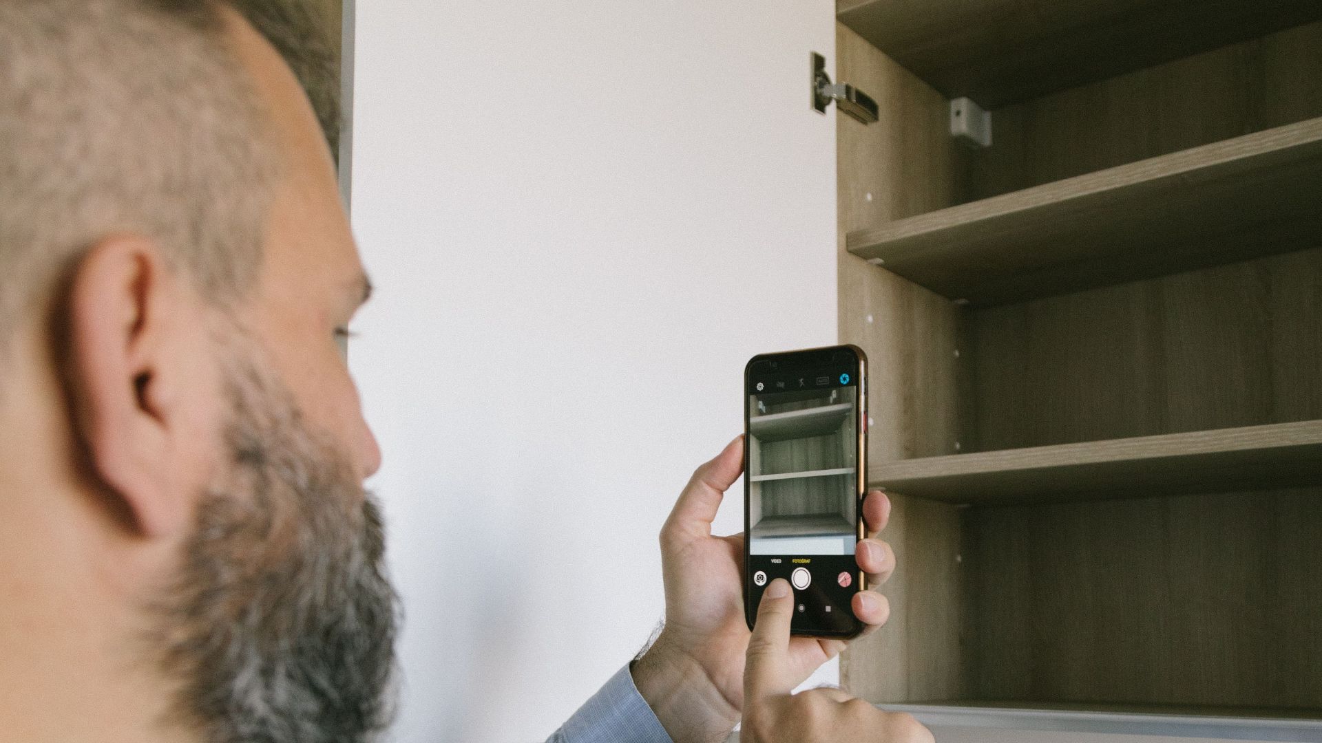 A bearded man uses a smartphone to photograph an opened kitchen cabinet.