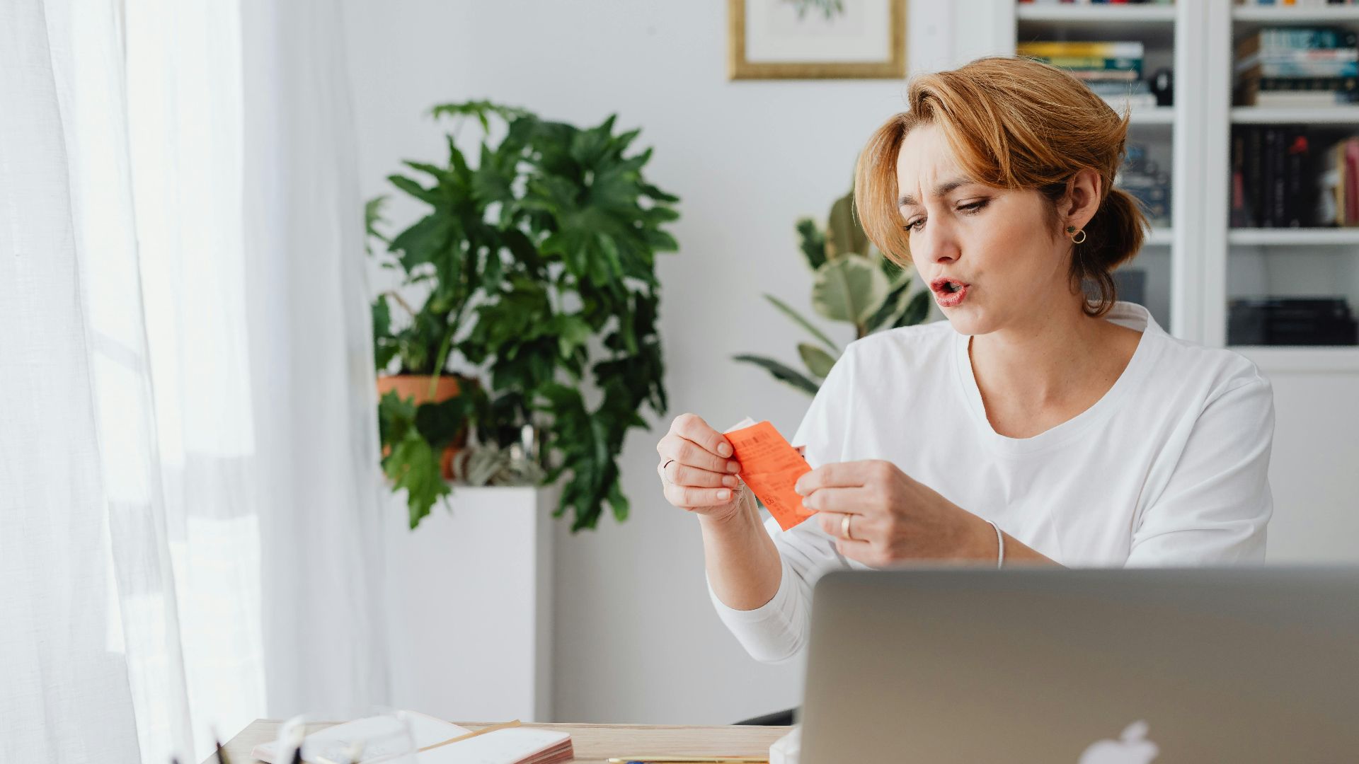 Woman reviewing bills at home desk with laptop and plants, managing personal finances.