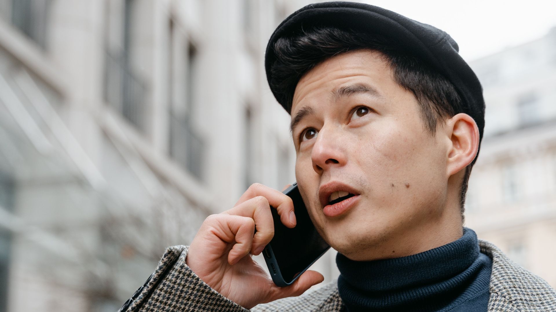 Stylish man in a city talking on a smartphone outdoors, wearing a coat and hat.