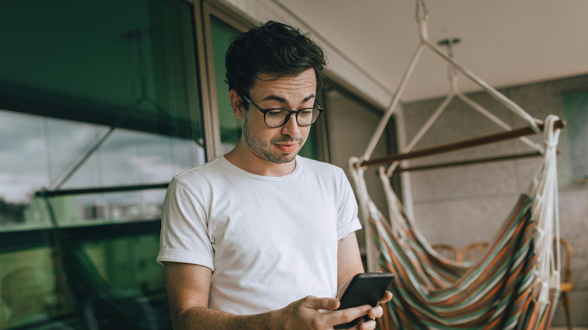 Young man in glasses using smartphone indoors with striped hammock.