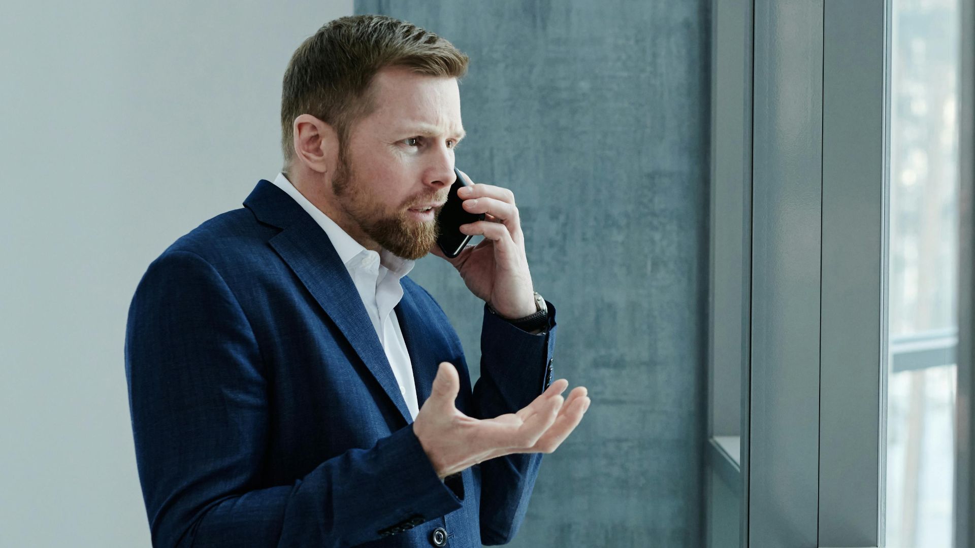 Businessman using smartphone while standing indoors by glass window, expressing concern.