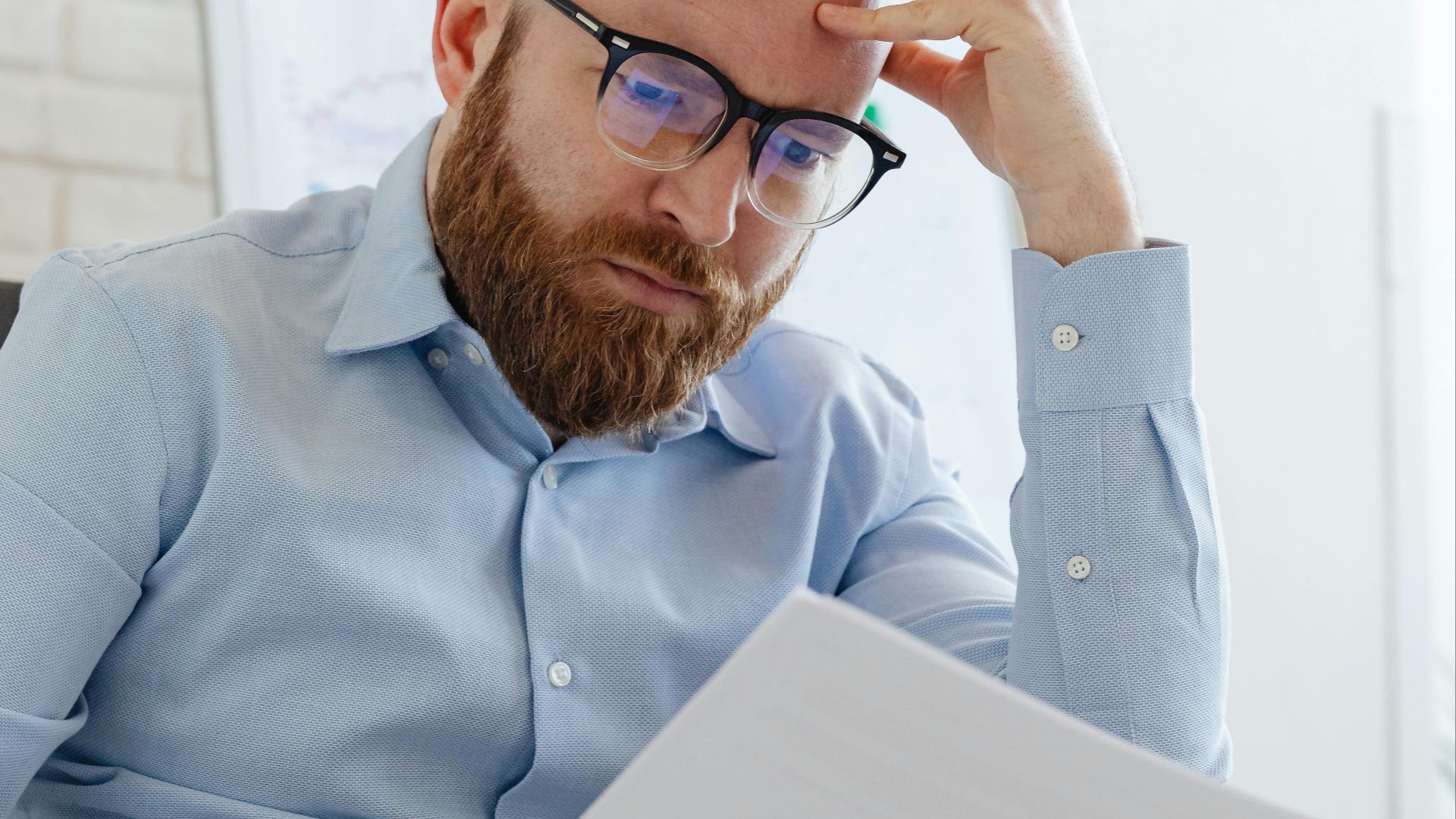 Caucasian businessman with beard and glasses reviewing documents in a modern office setting.