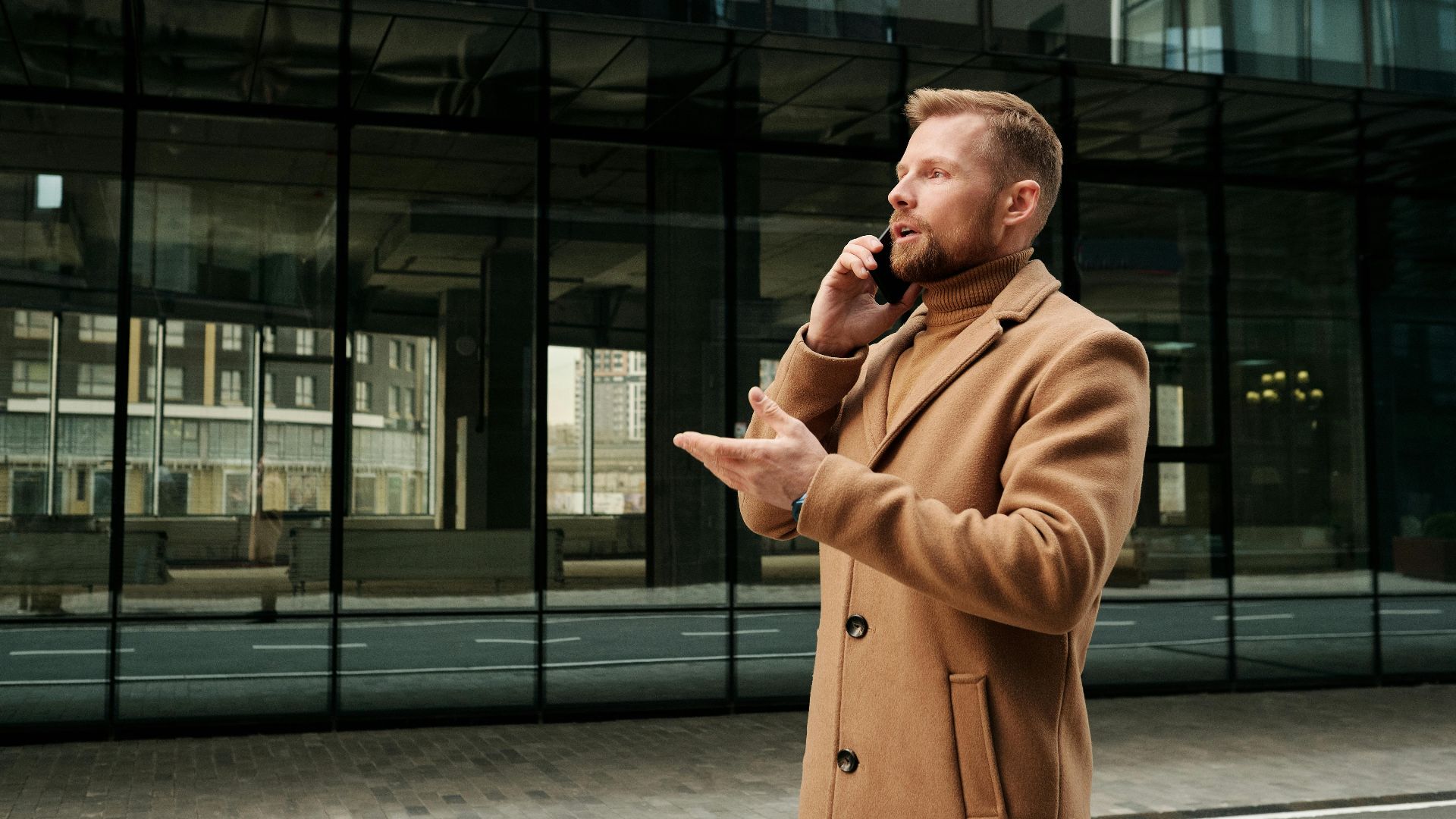 Confident businessman in a brown coat talks on phone outdoors in city.