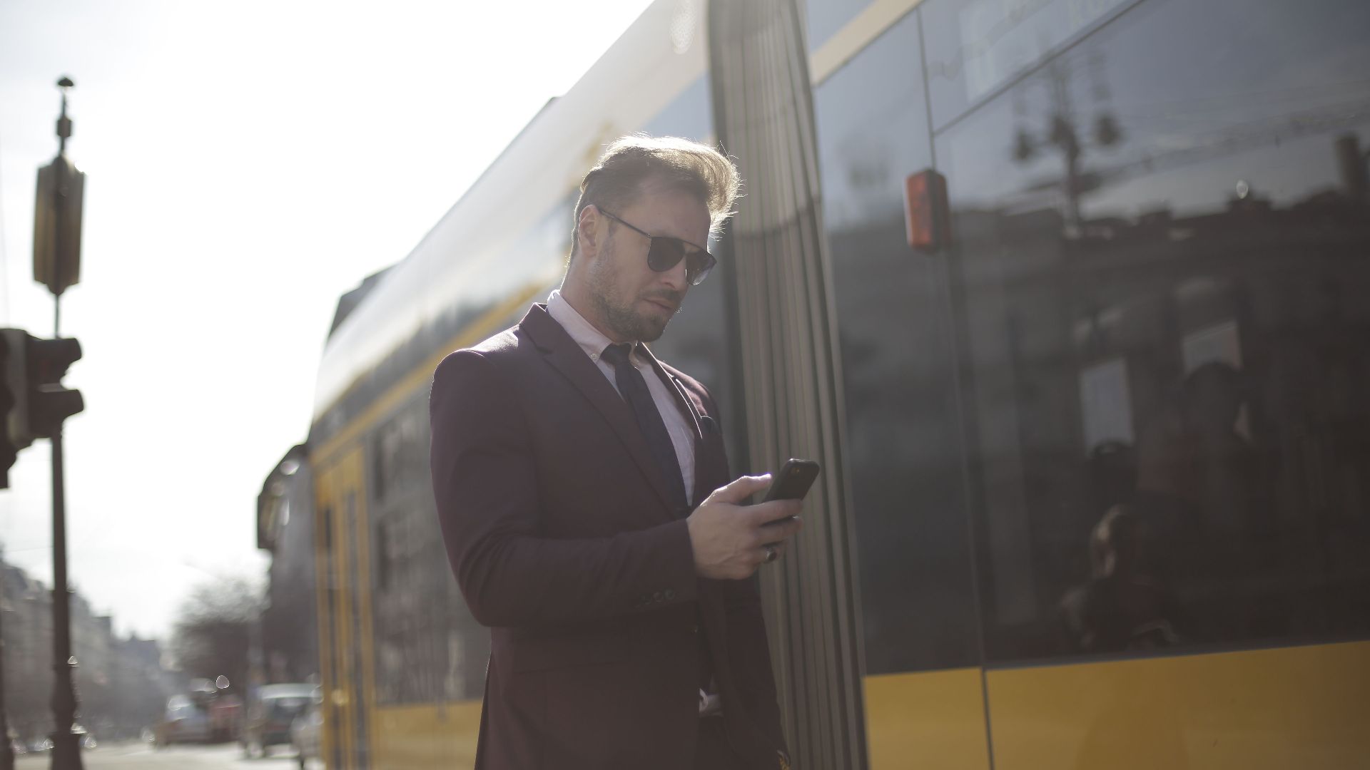 Business professional checks phone at a sunny train station, wearing a suit and sunglasses.