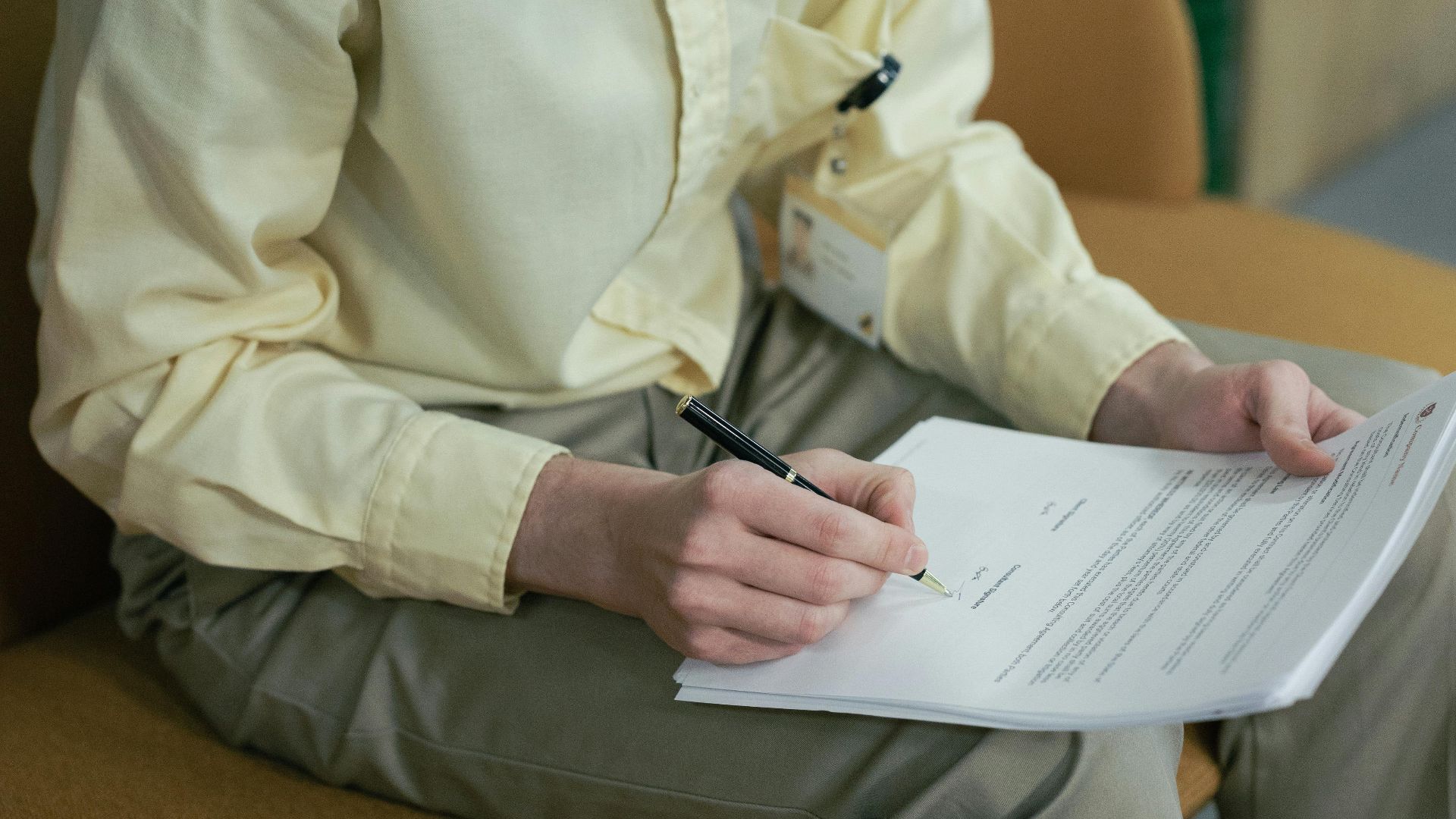 A young man with curly hair and glasses signs papers while sitting indoors.