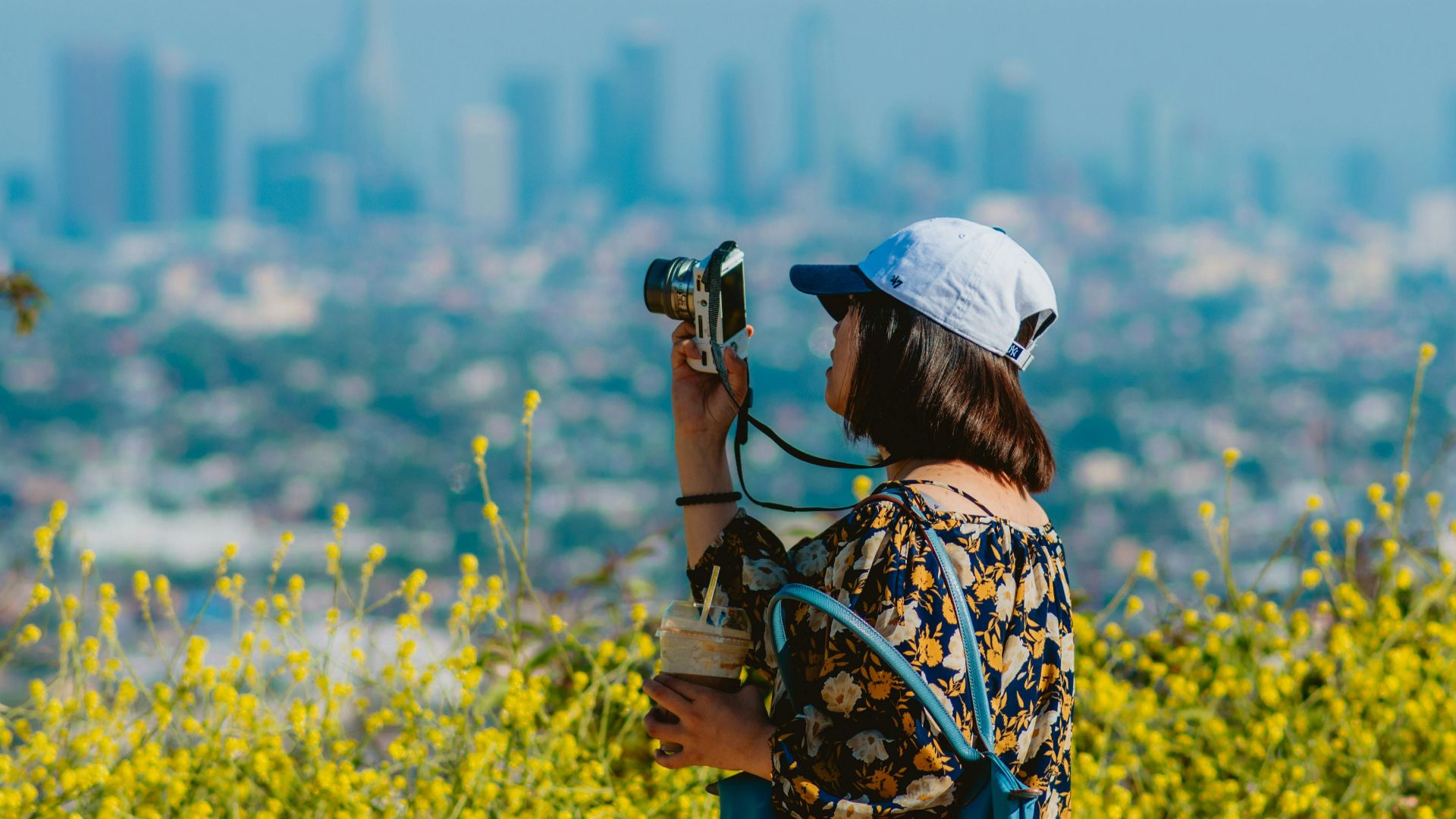 A woman in a floral dress photographs a city skyline with yellow flowers in the foreground.