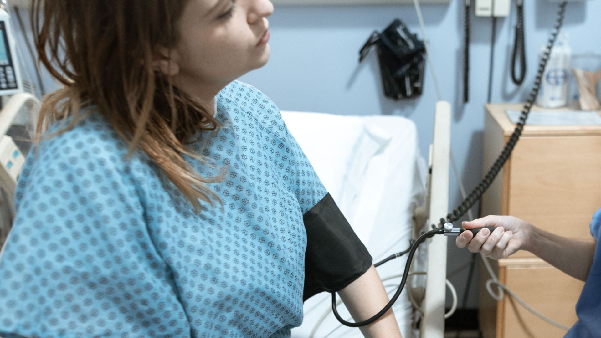 A nurse in blue scrubs checks the blood pressure of a female patient in a hospital room.