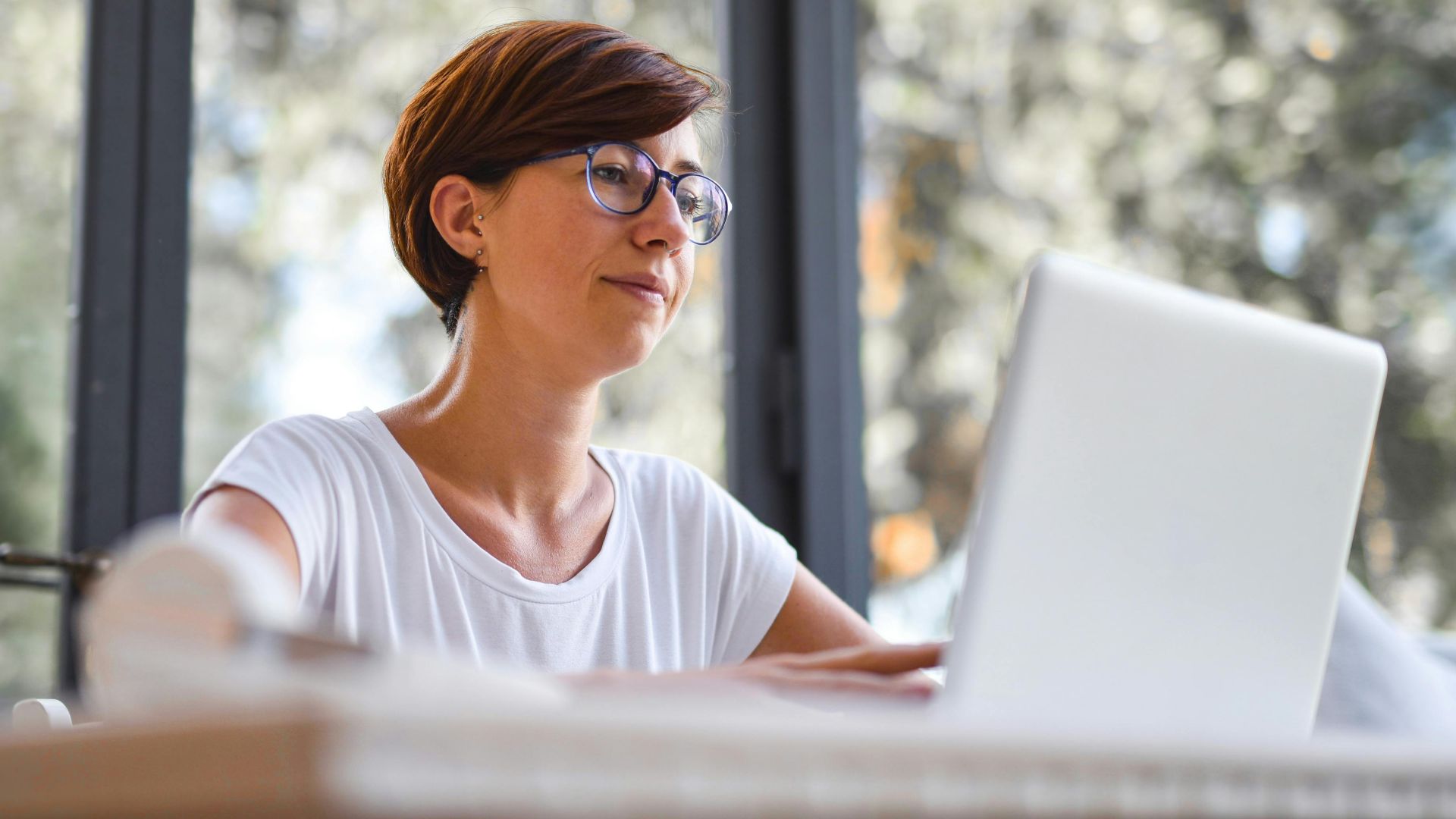 Woman with glasses using laptop in a bright home office, enjoying a peaceful work environment with natural light.