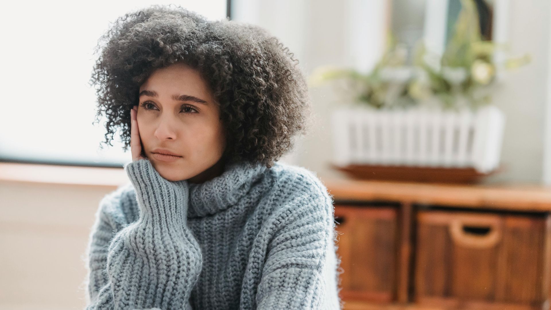 Discontent African American female leaning on hand and looking away while sitting in light room near green plant on blurred background