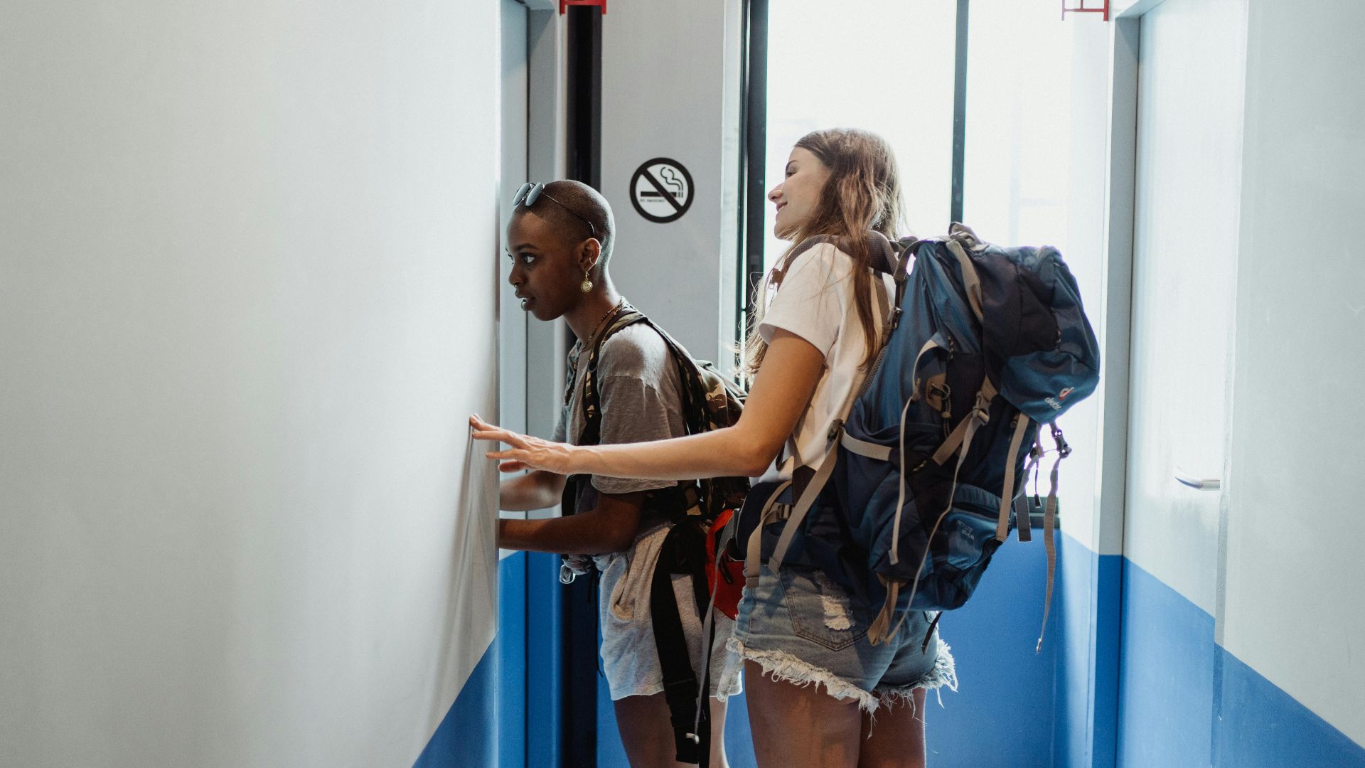 Two women with backpacks entering a hostel room, ready for travel.