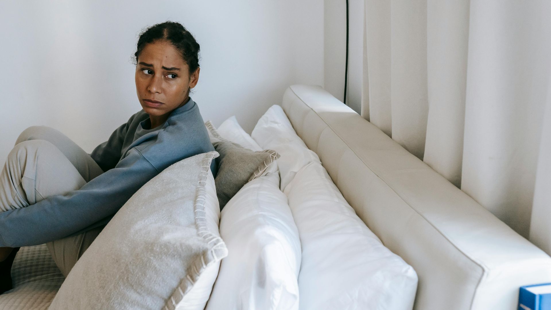 Sad young African American woman spending time on white bed and thinking in casual outfit while looking away at home