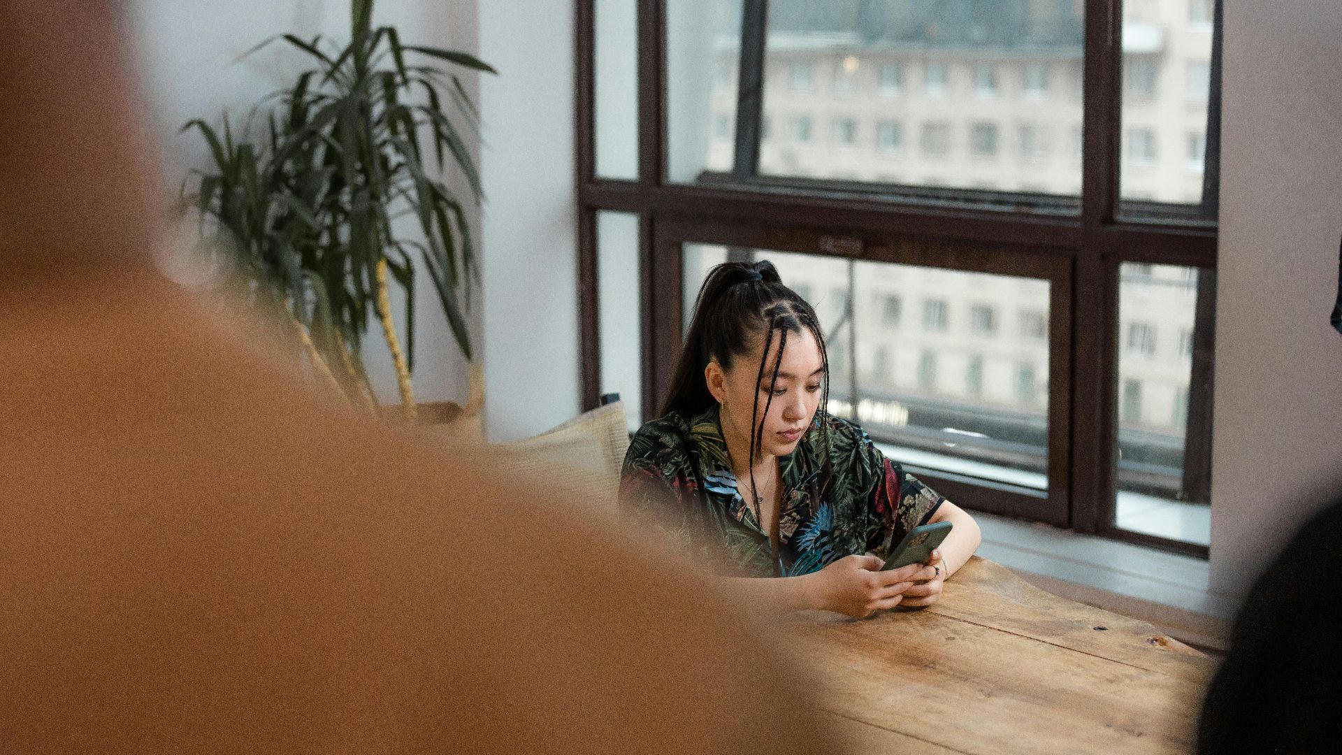 Young woman engrossed with a smartphone while sitting indoors by a large window.