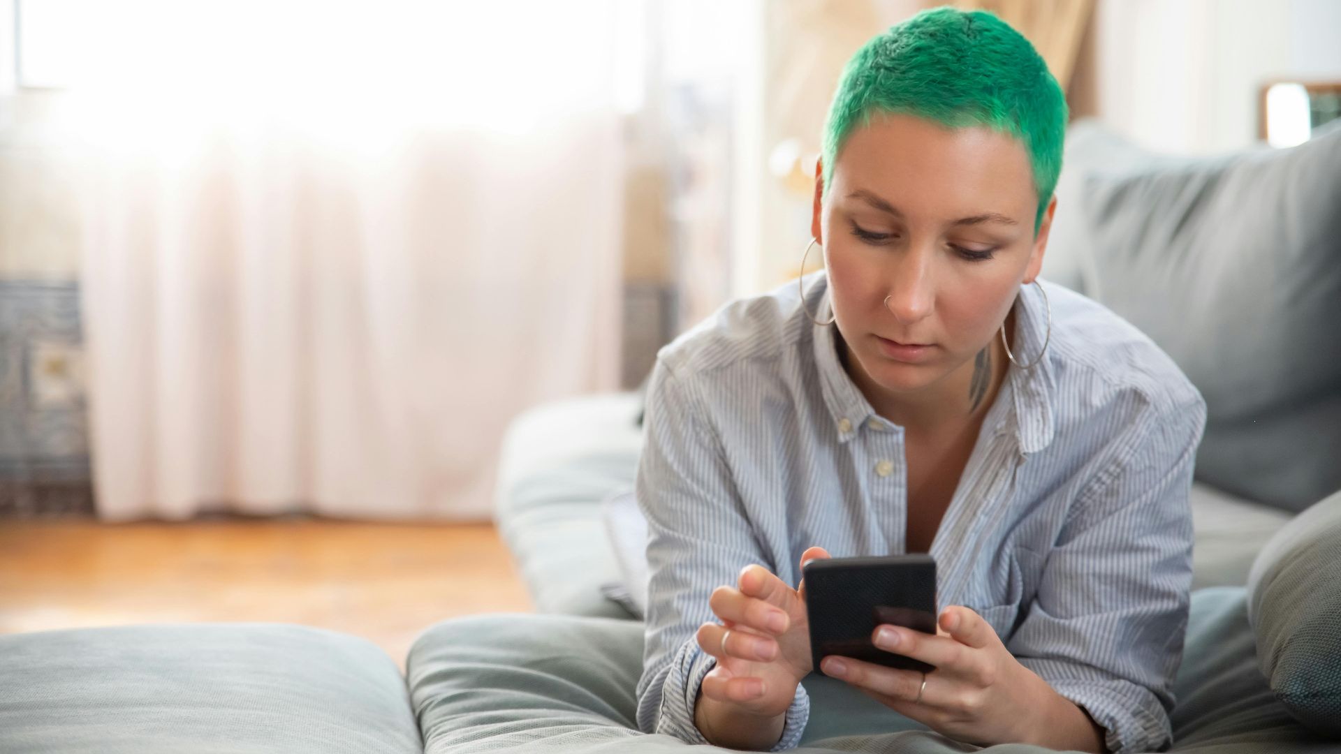 Woman with vibrant green hair lying on sofa using smartphone indoors.