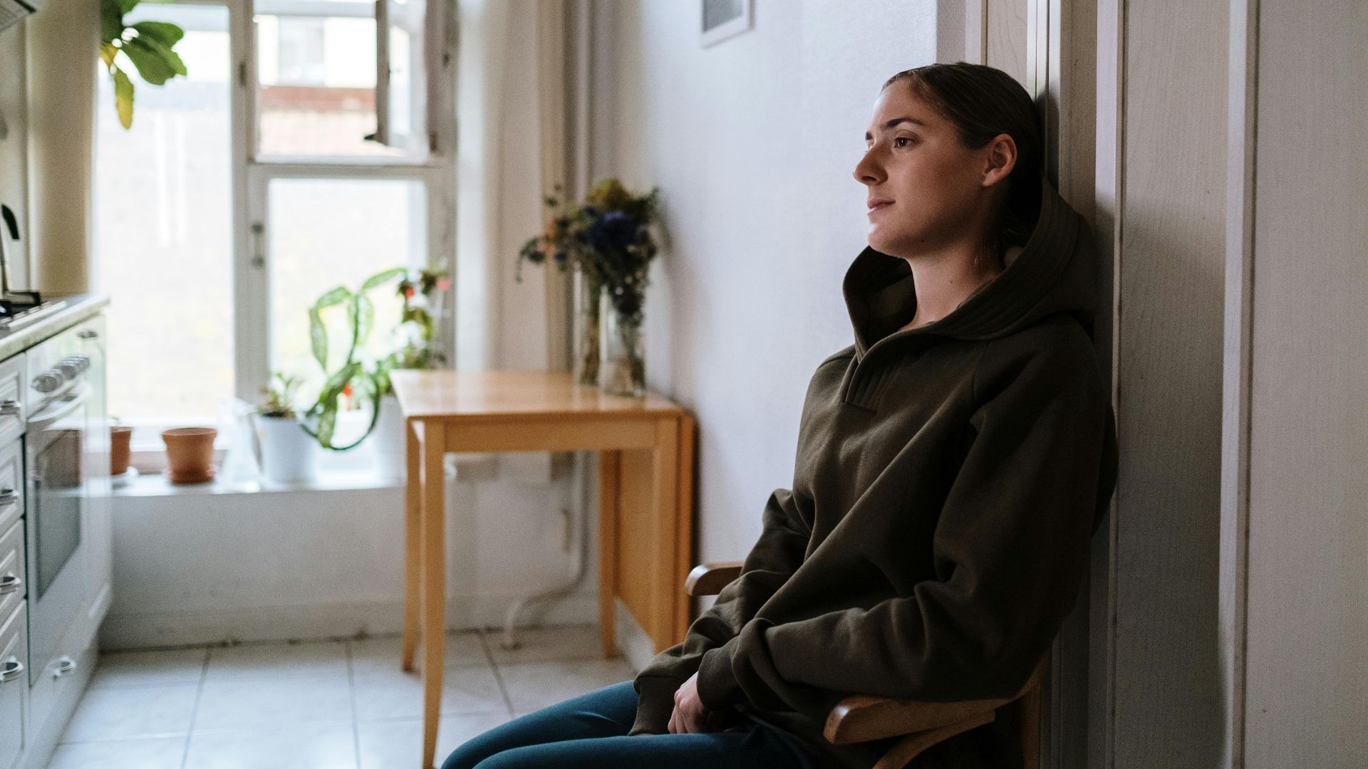 Pensive woman in a hoodie sitting indoors by a window, capturing a moment of reflection.