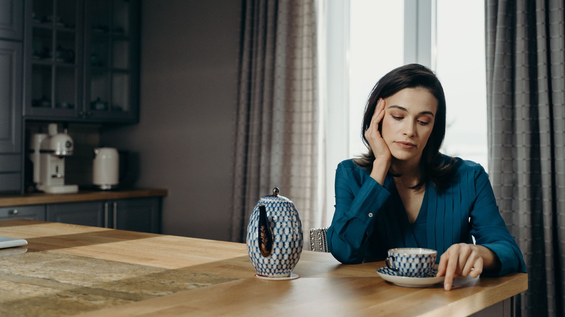 Woman in a blue blouse relaxing with coffee at a wooden table in a cozy dining room.