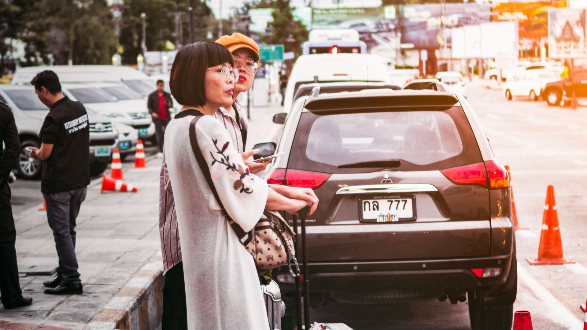 Two women with luggage waiting by a parked car on a busy street in Thailand.