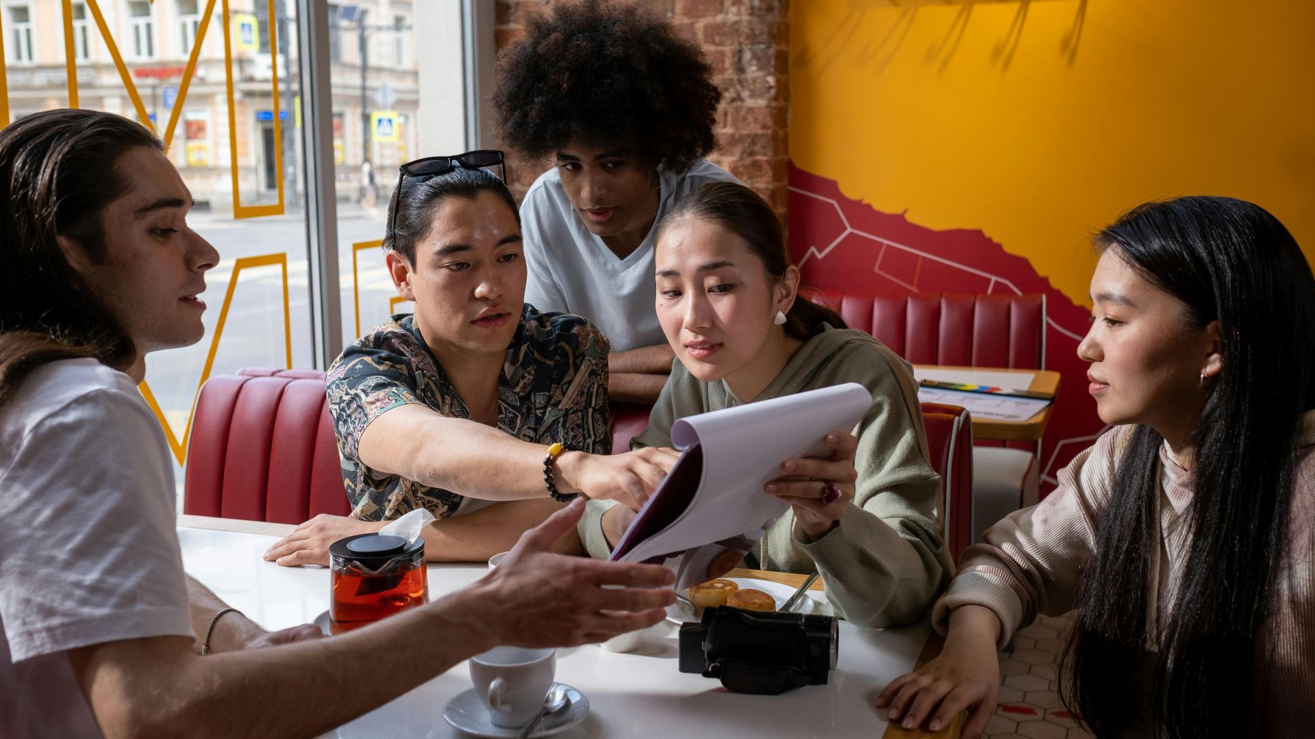 A multicultural group of young adults engaged in conversation around a table in a cafe, focused on a document.