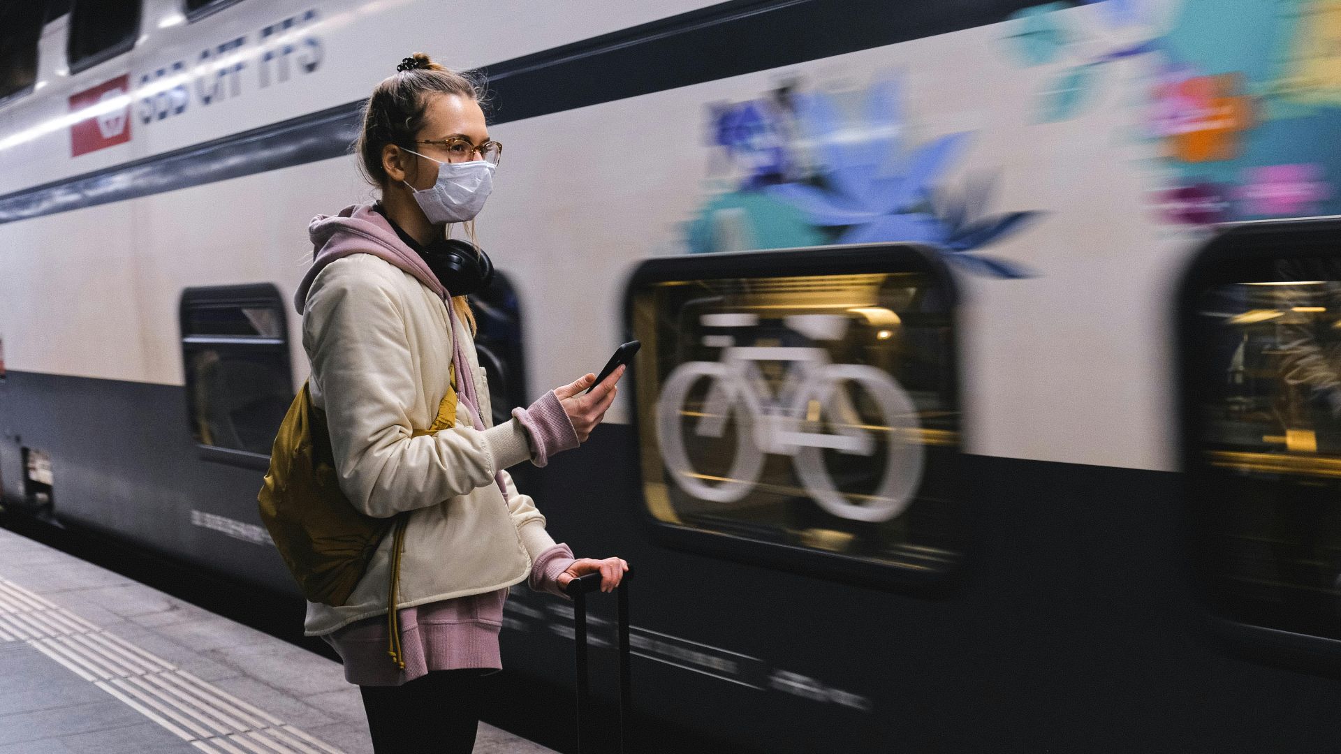 Woman wearing mask stands with luggage using cellphone in subway station.