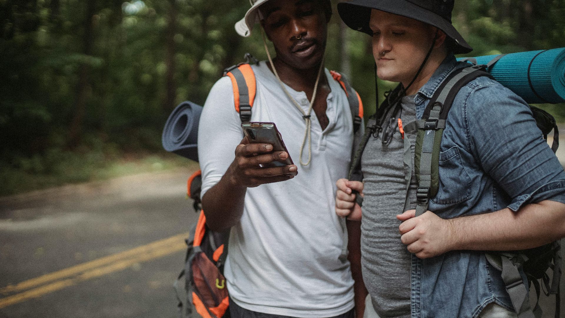 Crop multiracial travelers with backpacks sharing cellphone while talking on countryside roadway in summer