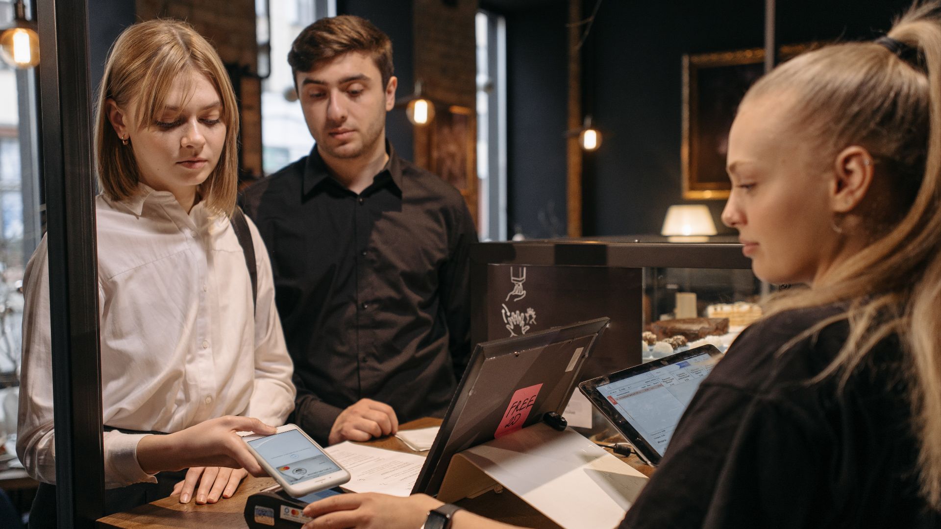 Two customers making a contactless payment at a café counter using a mobile phone.