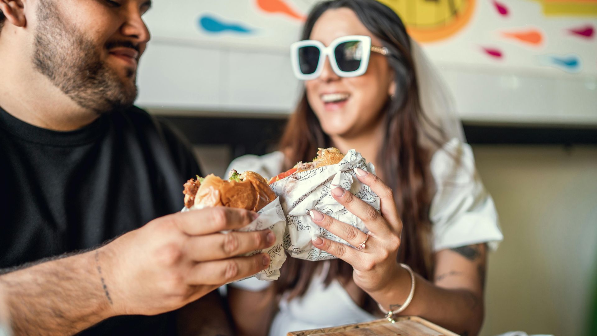 Two friends laughing while sharing a burger meal indoors. Fun and relaxation.