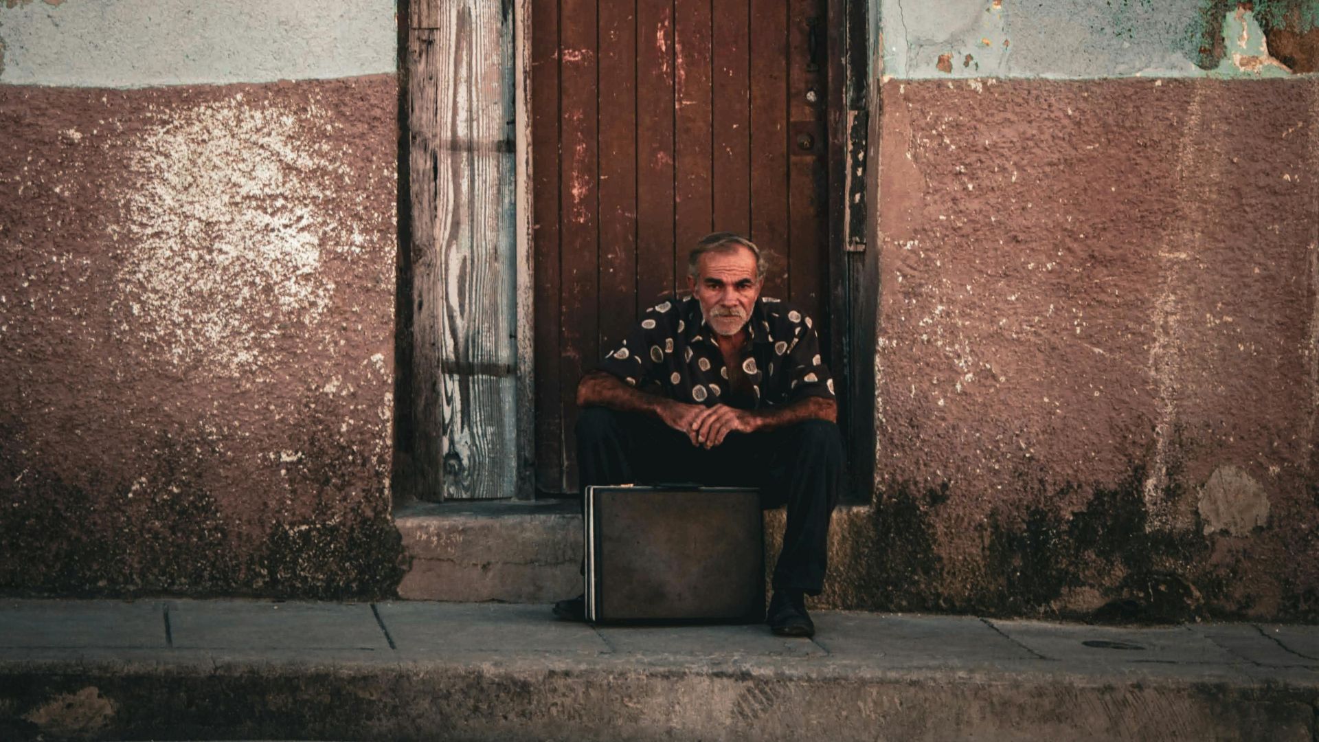 An elderly man sits next to a weathered door in a rustic urban setting, conveying quiet reflection.