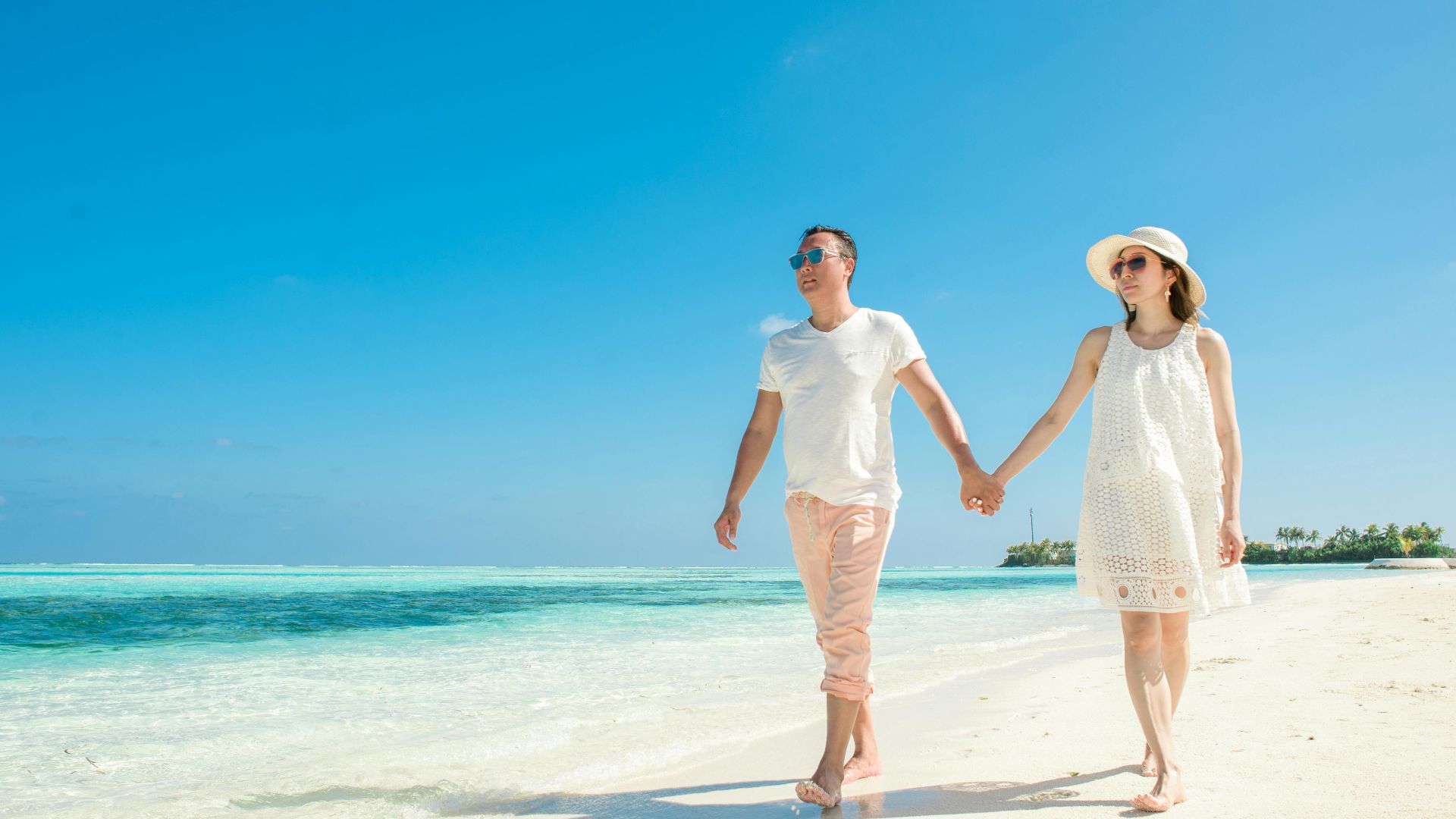A couple holding hands and walking barefoot on a sandy beach under a clear blue sky.