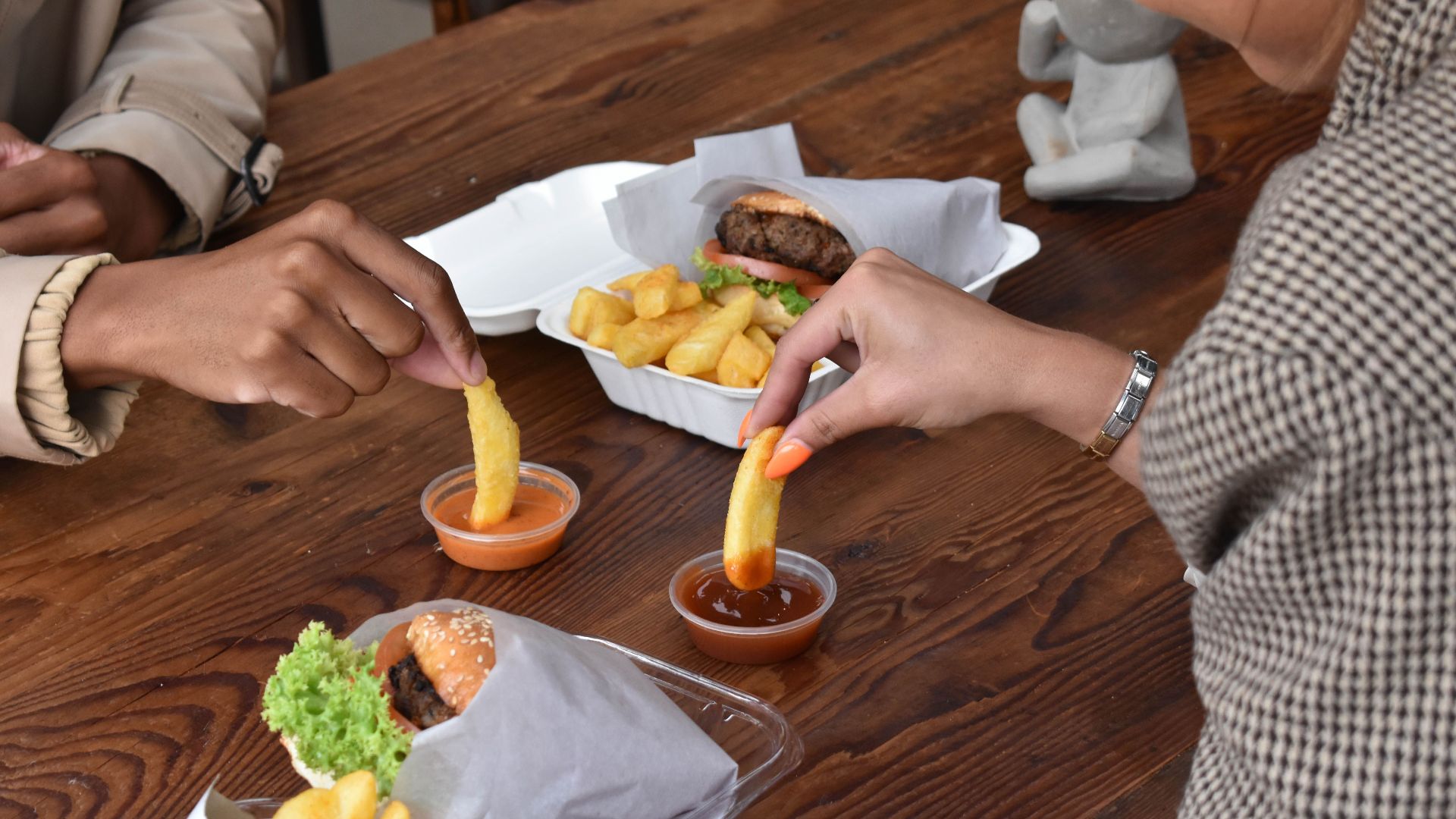 Friends sharing a meal of hamburgers and fries, dipping in sauces at a wooden table.