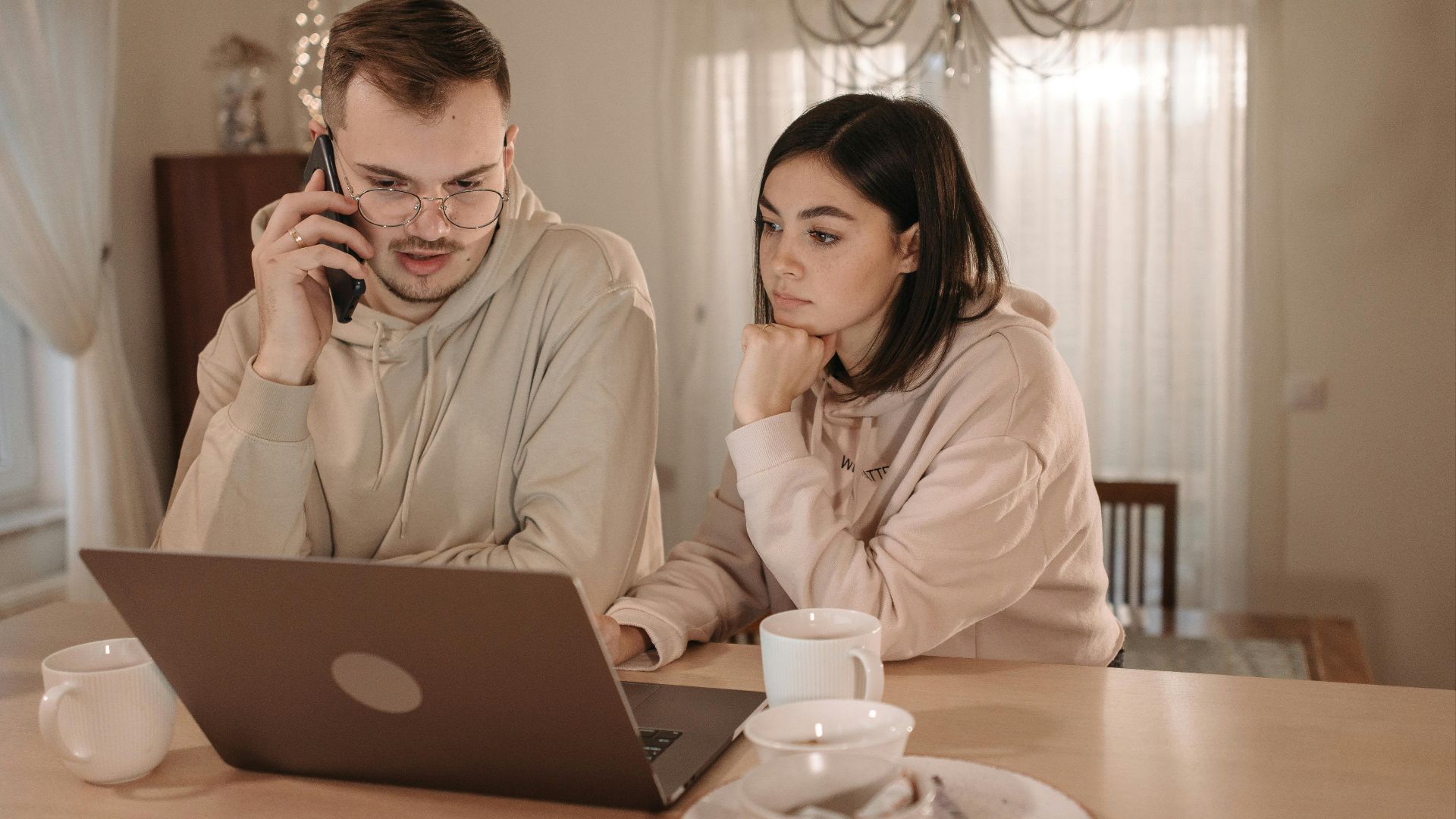 A couple in hoodies focused on work using a laptop and smartphone at home.