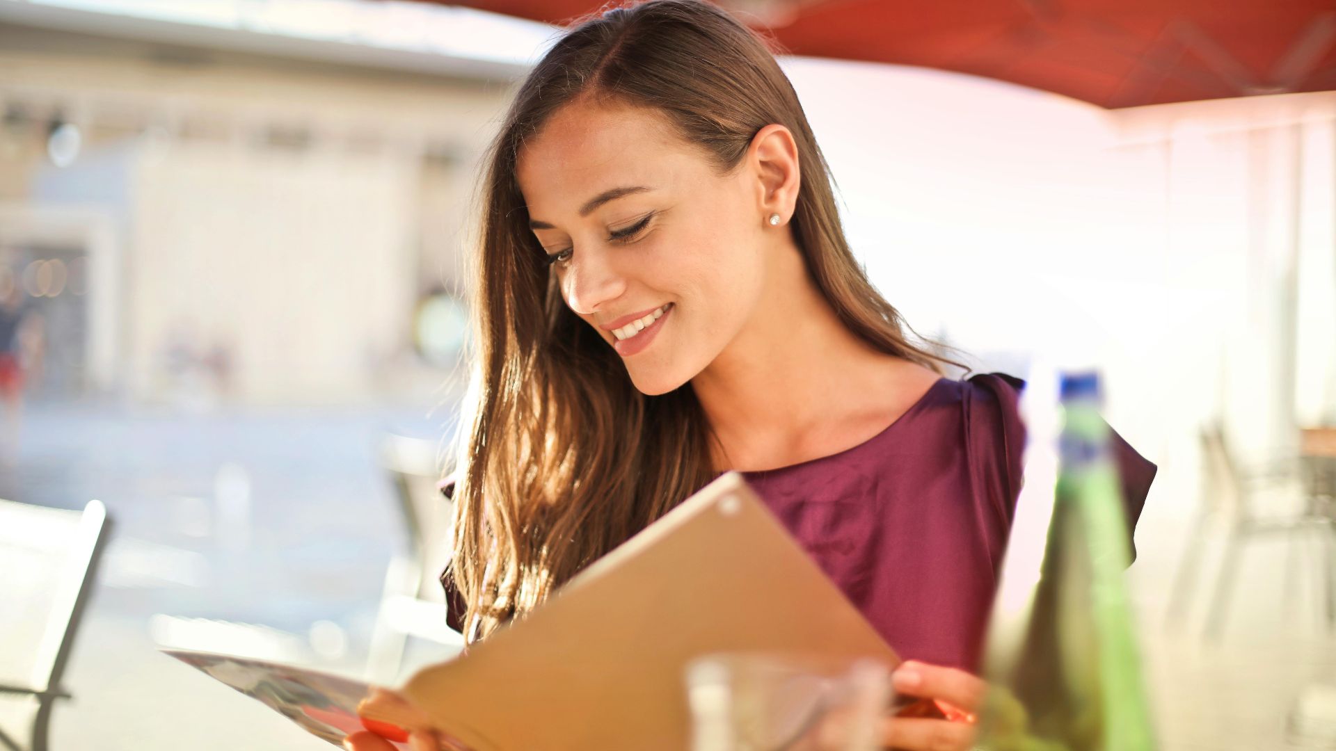A woman smiles as she reads a menu at an outdoor restaurant, enjoying a sunny day.
