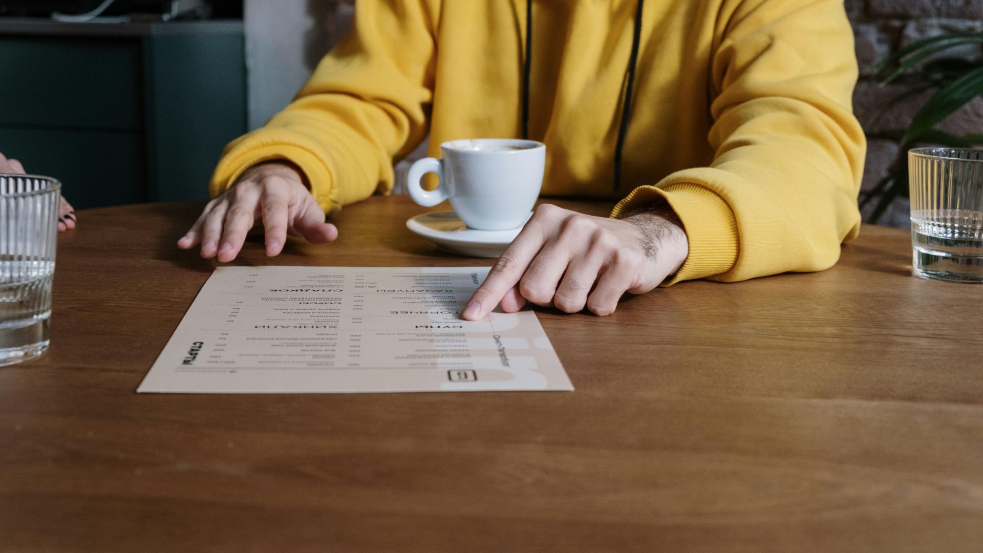 A person wearing a yellow hoodie points to a menu in a cozy cafe setting.