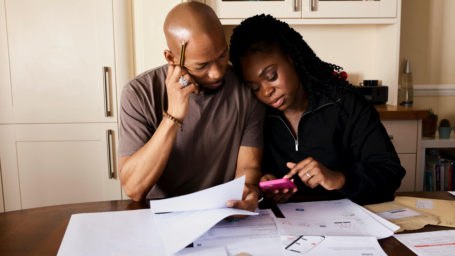 A couple sits at a table managing domestic finances, evaluating documents and using a smartphone.