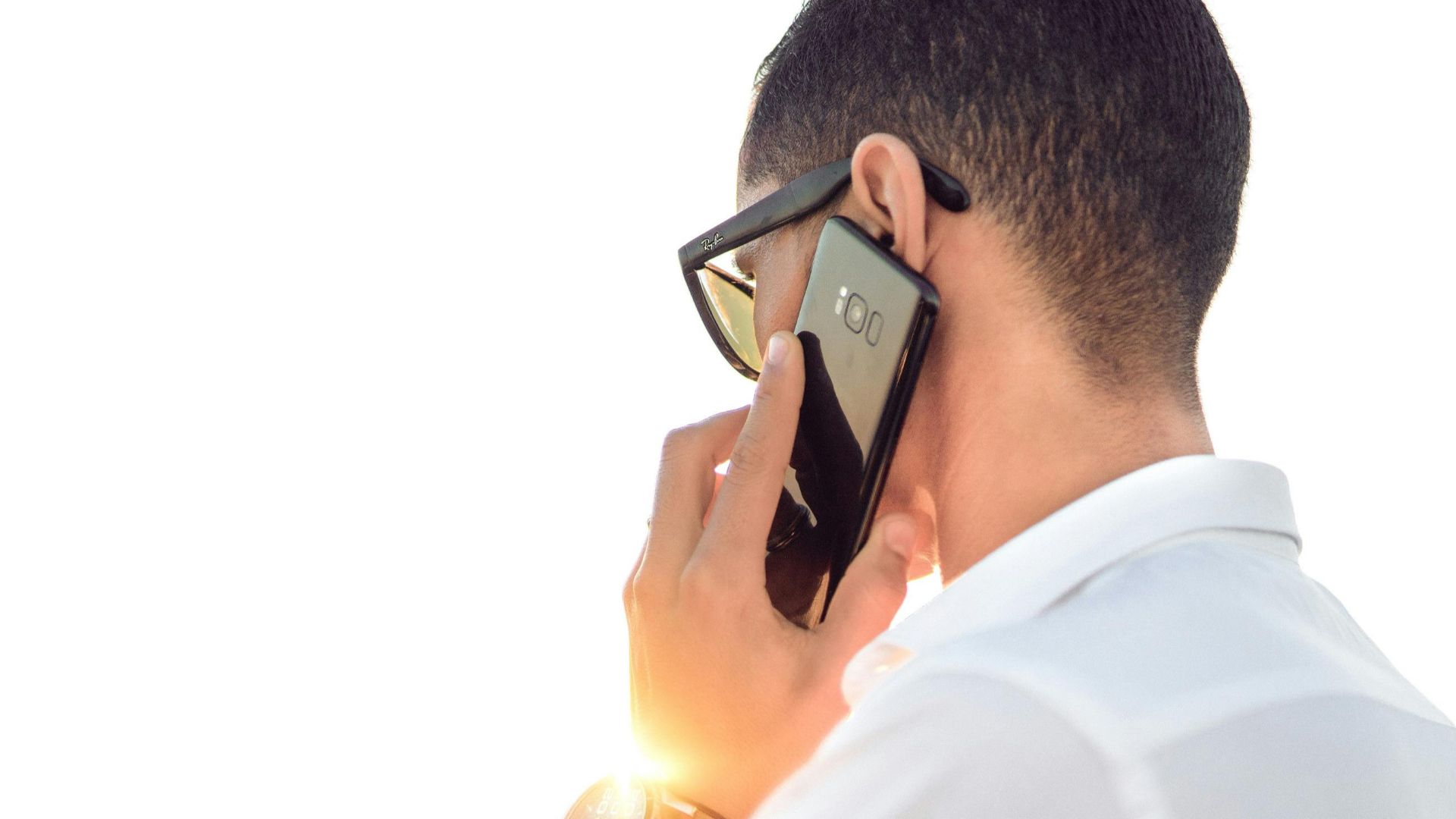 Man outdoors in Morocco talking on smartphone, wearing sunglasses and white shirt, backlit by sunset.