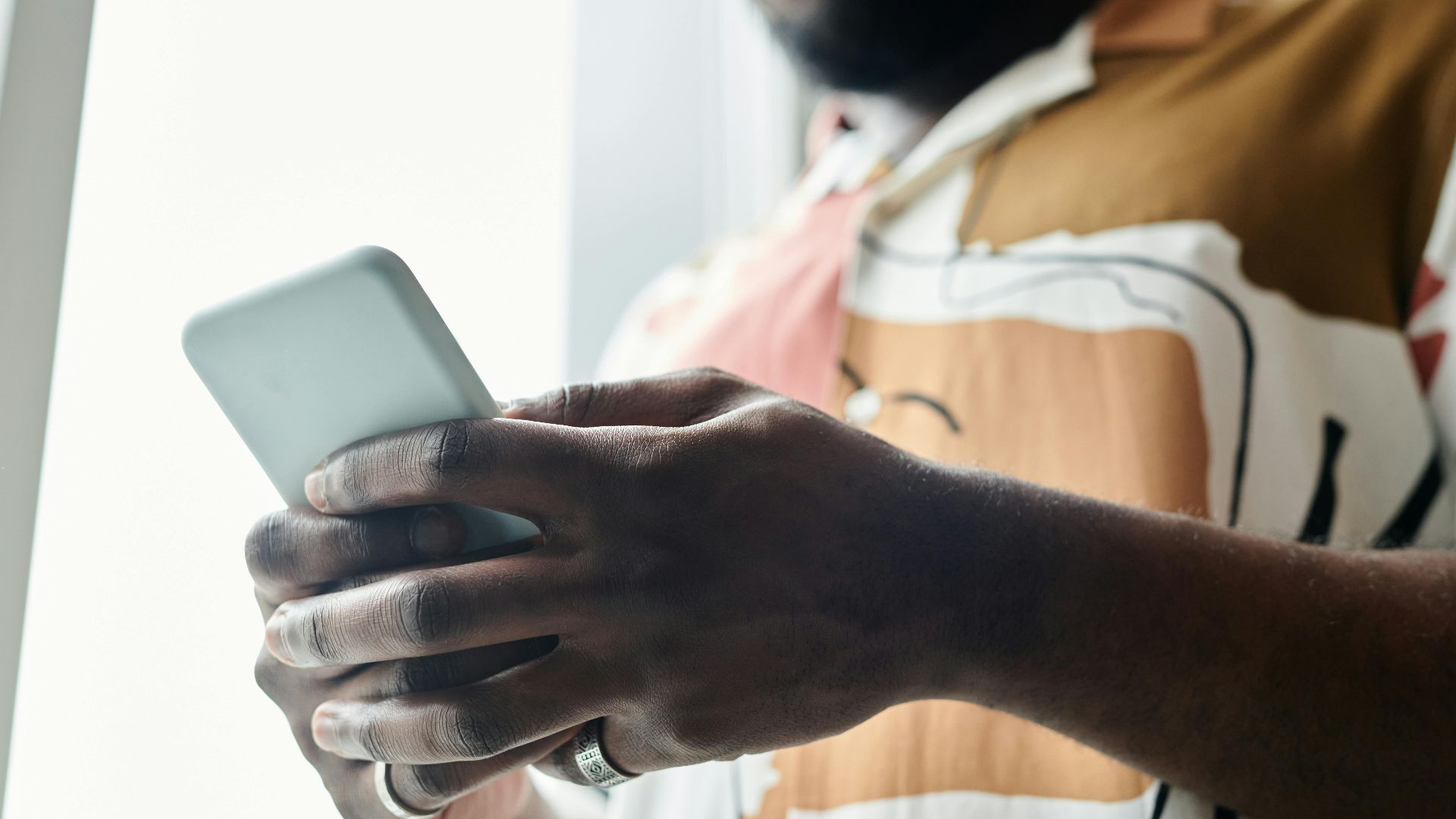 A close-up of a man using a smartphone by a window, featuring a modern patterned shirt.