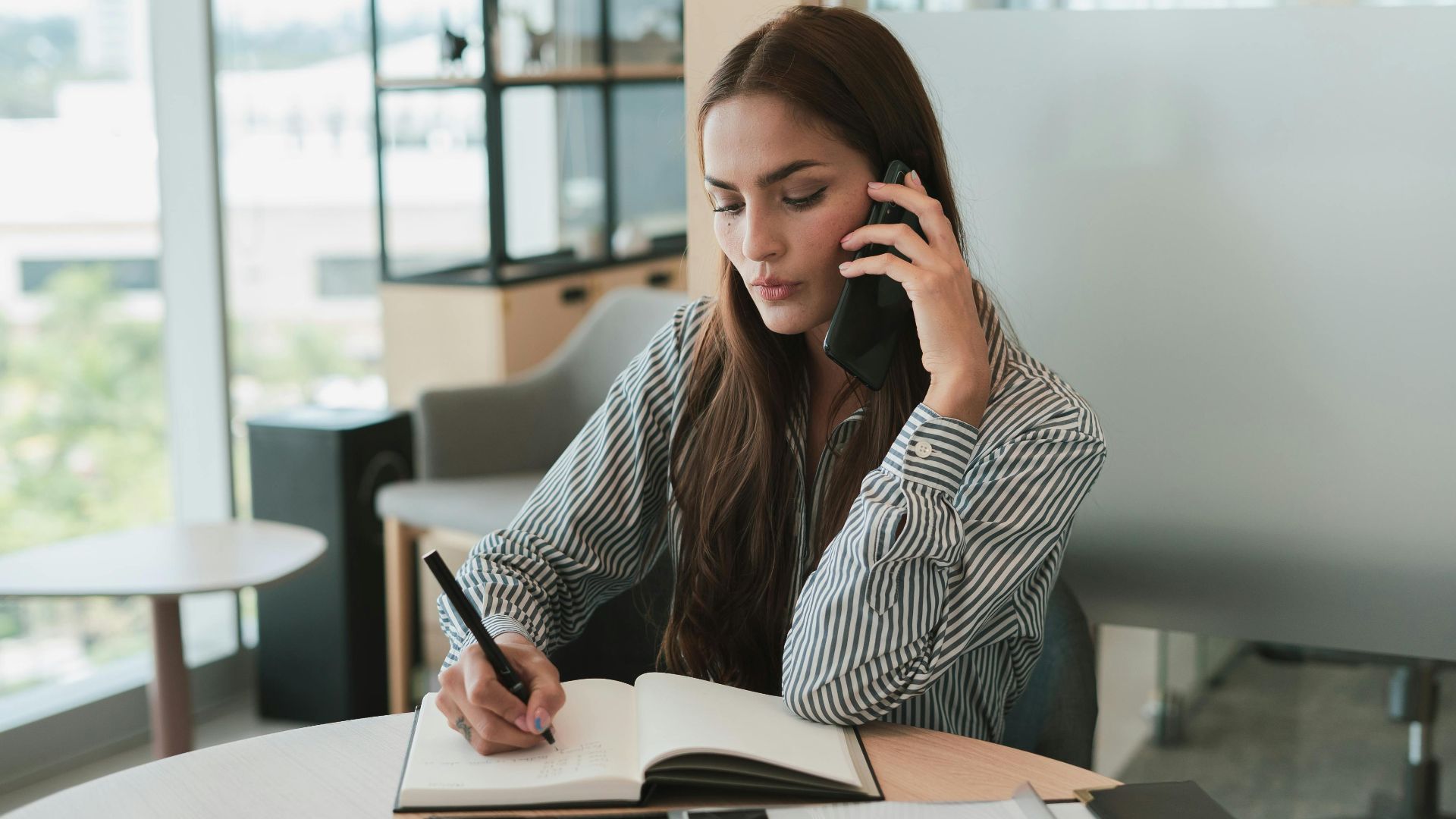 Young businesswoman on phone, jotting notes at stylish office desk.