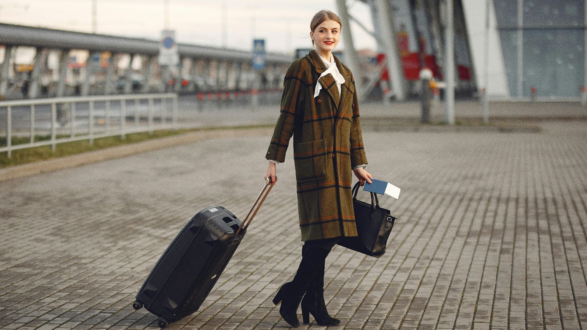 Full body young female passenger in trendy plaid coat with suitcase strolling on pavement near modern building of ariport terminal taking bag and documents