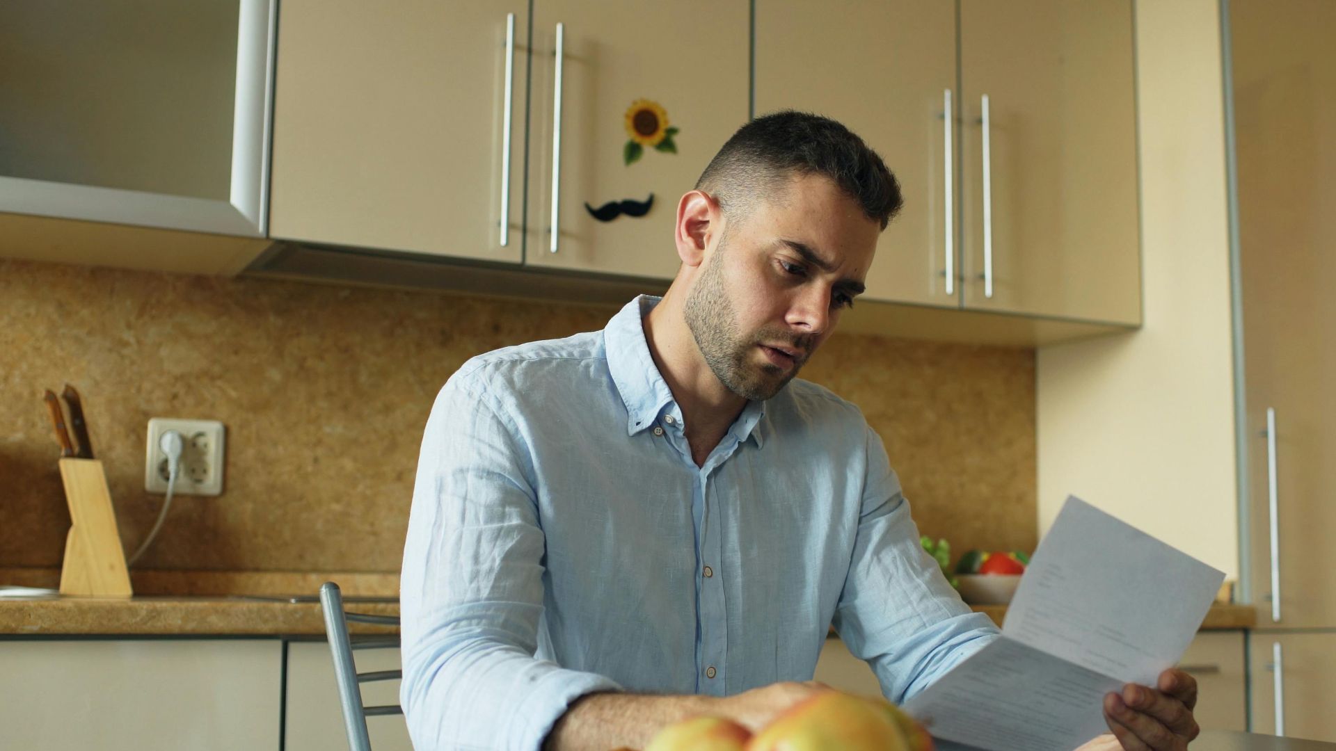 Man in a kitchen reading a document, apples on table.