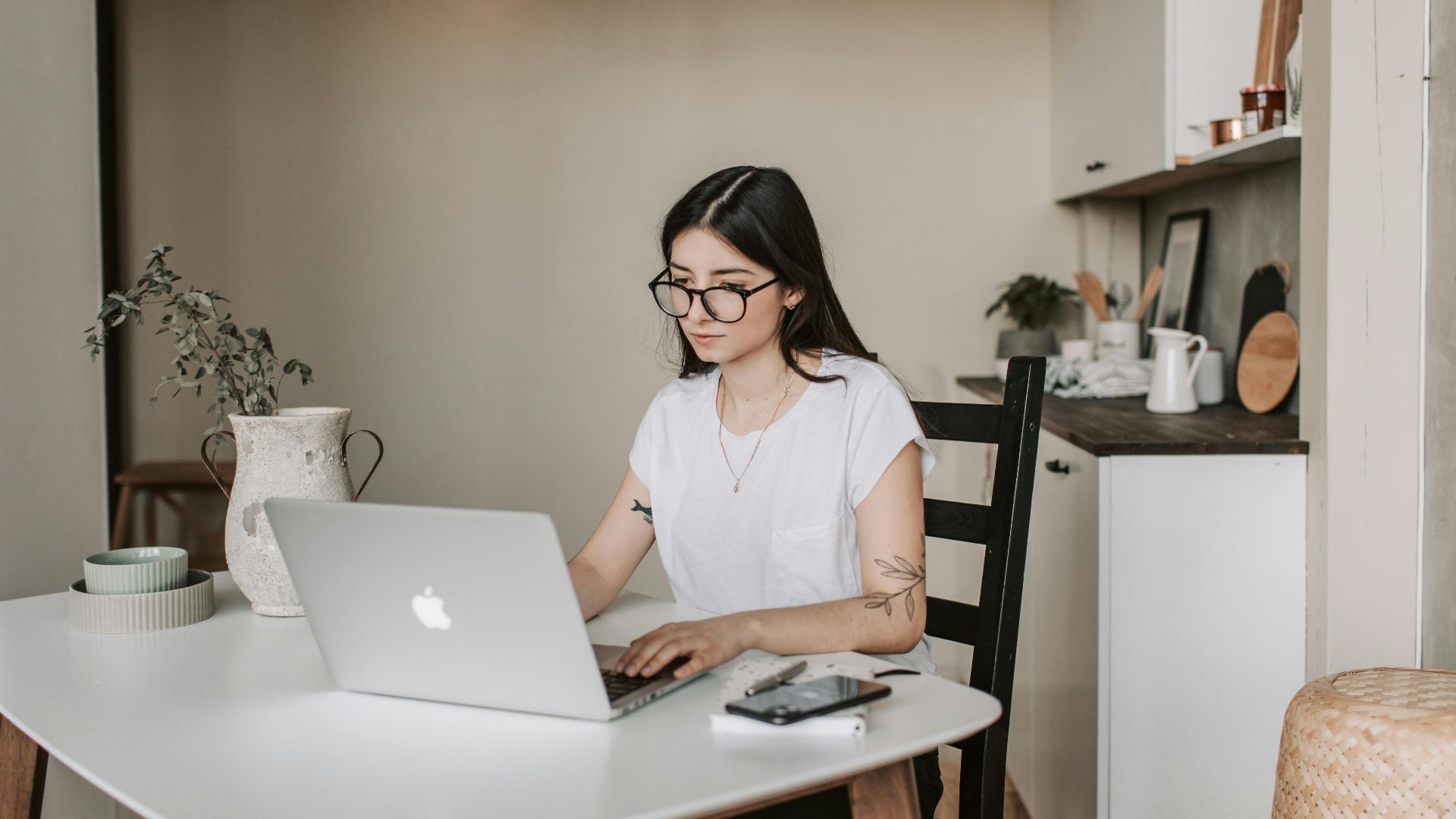Smart female freelancer with tattooed arms wearing eyeglasses working on remote project browsing netbook while sitting at table with smartphone and notepad in modern room interior
