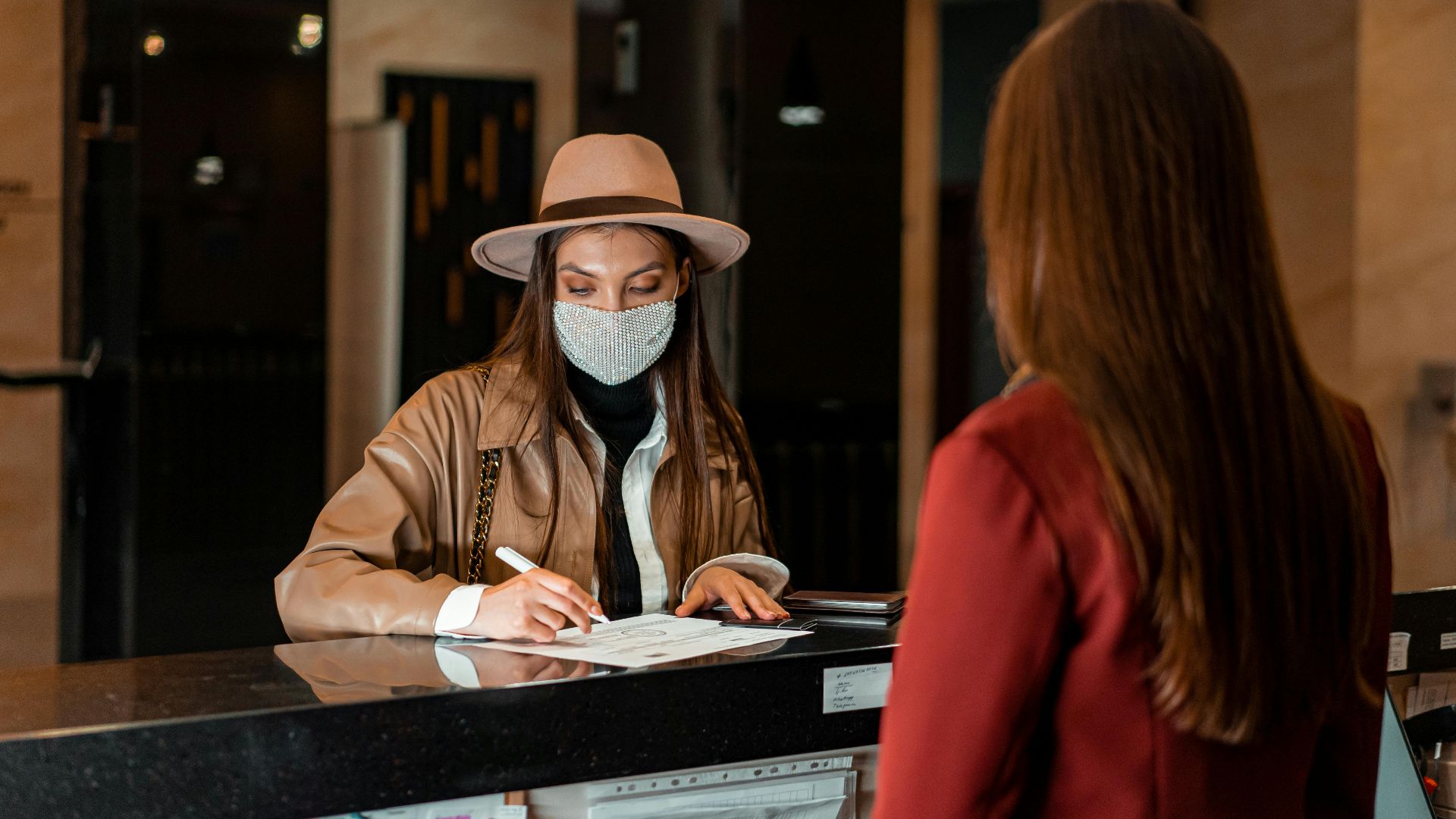 A woman in a hat and face mask checks in at a hotel reception, interacting with a receptionist.