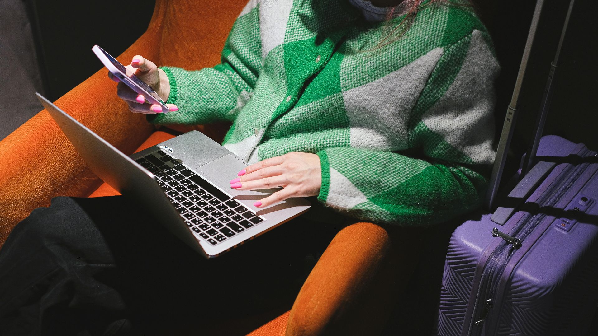 Woman in a knitted sweater working remotely on laptop with a smartphone, sitting in an armchair.