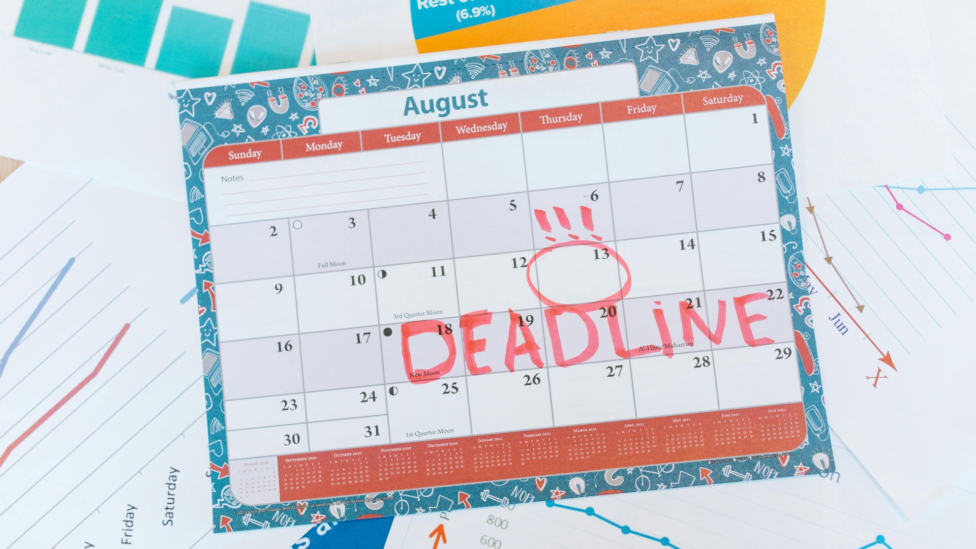 Vibrant August calendar on a desk with deadline marked in red, surrounded by graphs and charts.