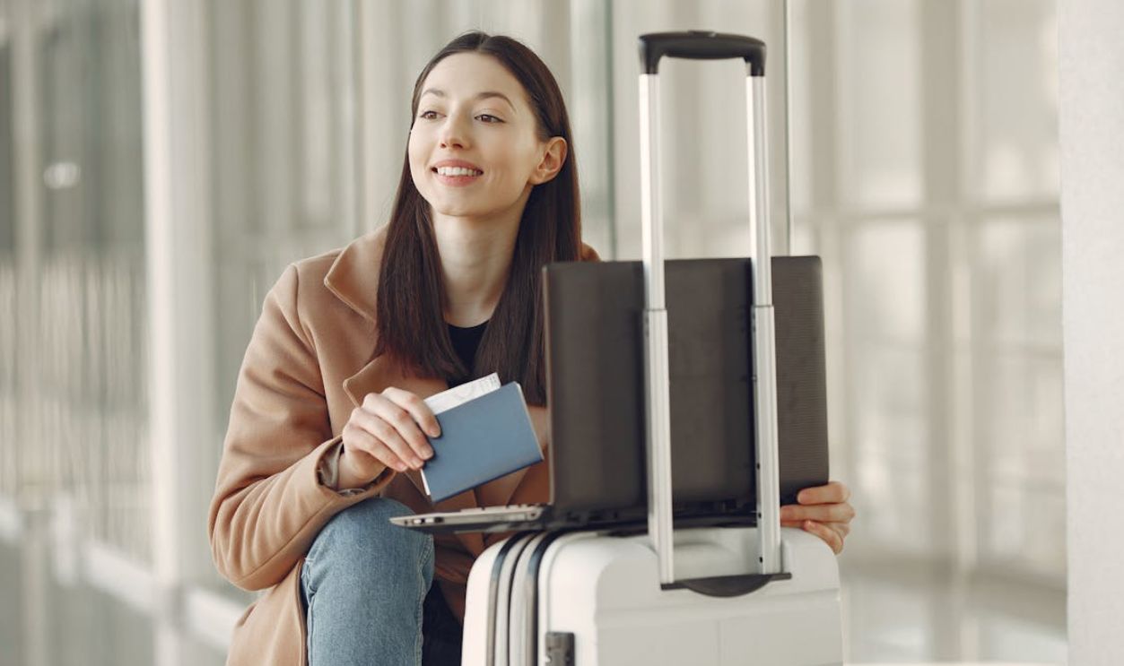 A woman sitting with her laptop and luggage, holding a passport, waiting in an airport terminal.
