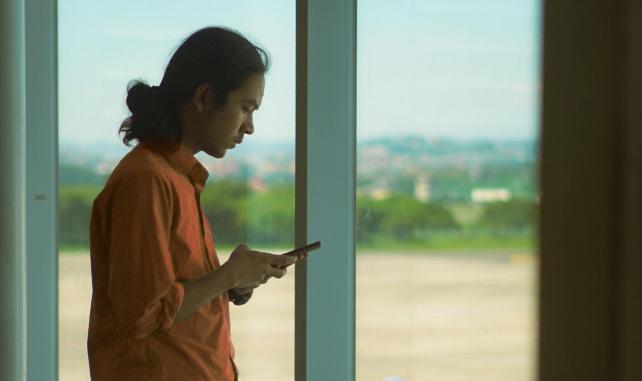 Man in orange shirt using a smartphone while standing at an airport window, overlooking the runway.