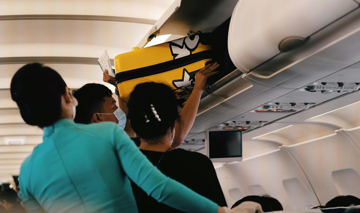 Passengers placing a yellow suitcase in the overhead bin of an airplane cabin. Interior with crew assistance.
