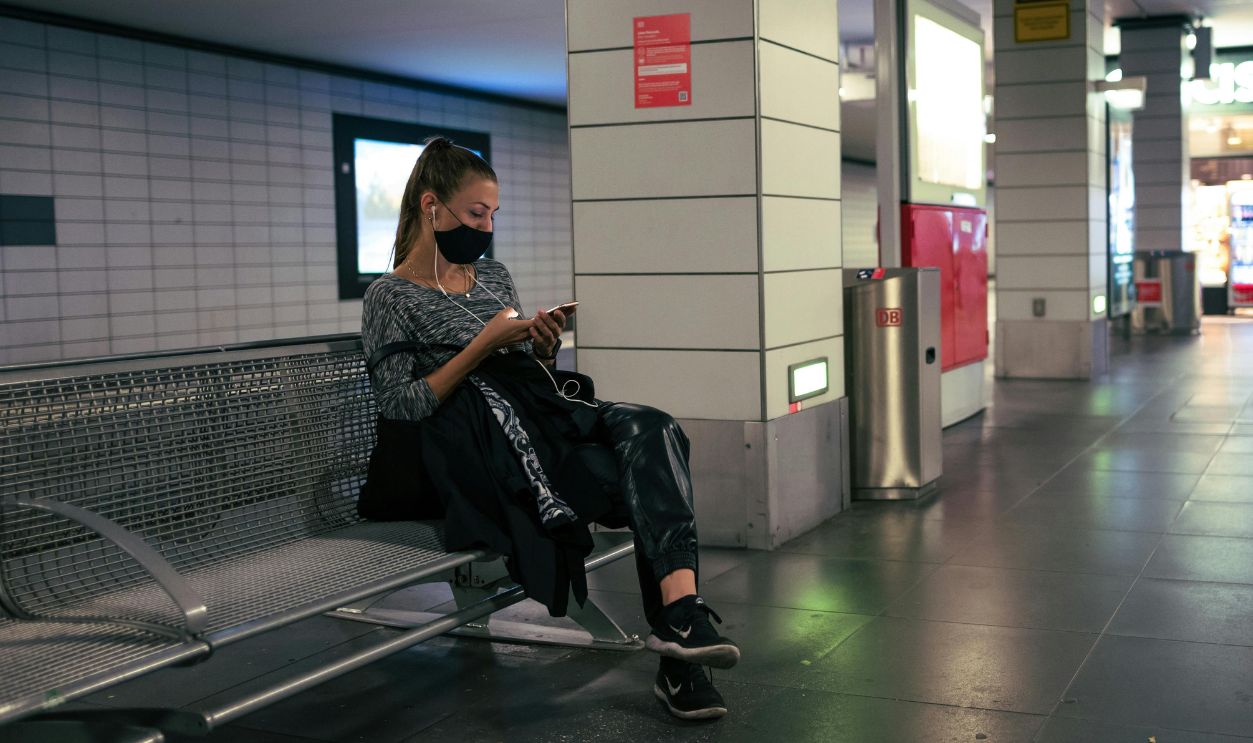 Woman sits on a bench in a subway station, wearing a face mask and using her phone.